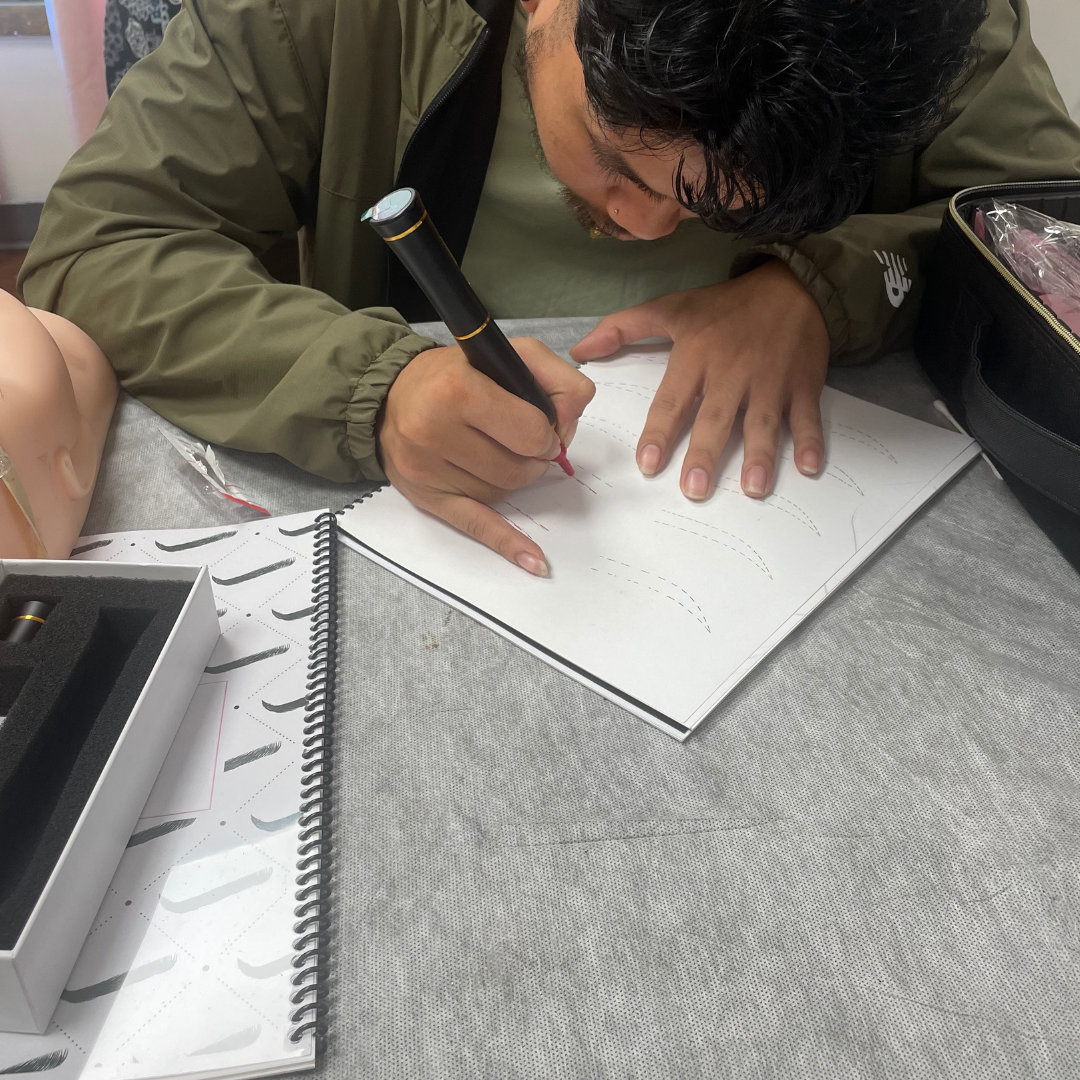 A person practicing calligraphy on a white notebook with a black marker, surrounded by practice sheets and supplies on a gray table.