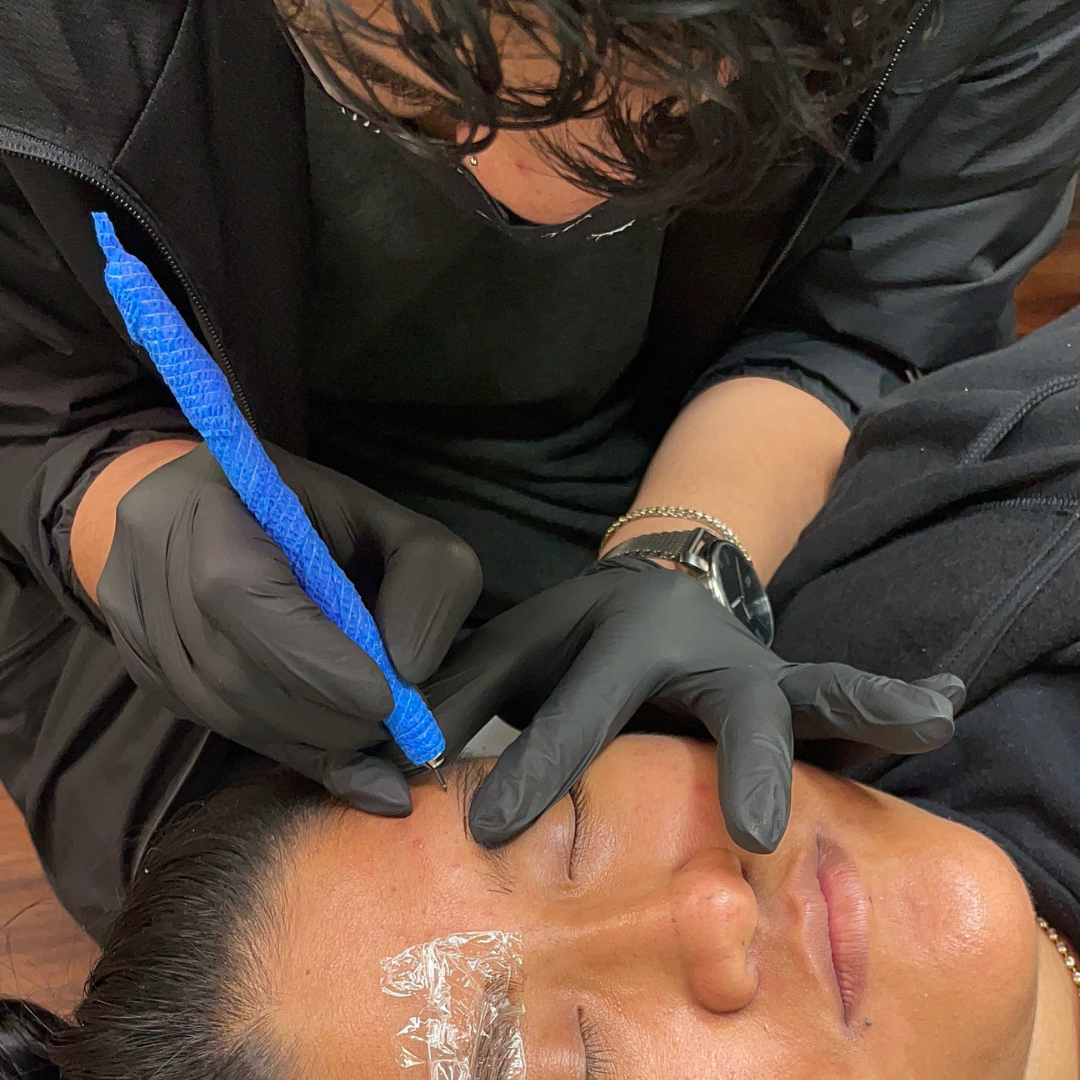 A person with dark hair receives a cosmetic tattoo procedure on their eyebrow from a professional wearing black gloves.