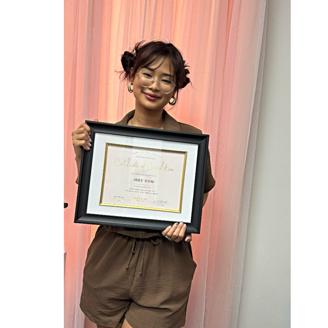 A woman with glasses and styled hair holding a framed certificate, smiling, in front of pink curtains.