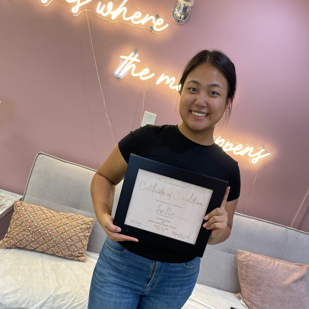 Young woman smiling and holding a framed certificate of completion in a pink room with a neon sign behind her that reads 'where the men's' and a gray sofa with pink pillows.