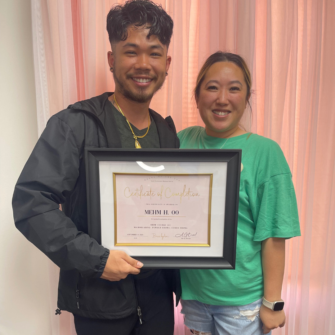 A man and woman smiling and holding a framed certificate of completion for a brow microblading course, indoors with pink curtains in the background.