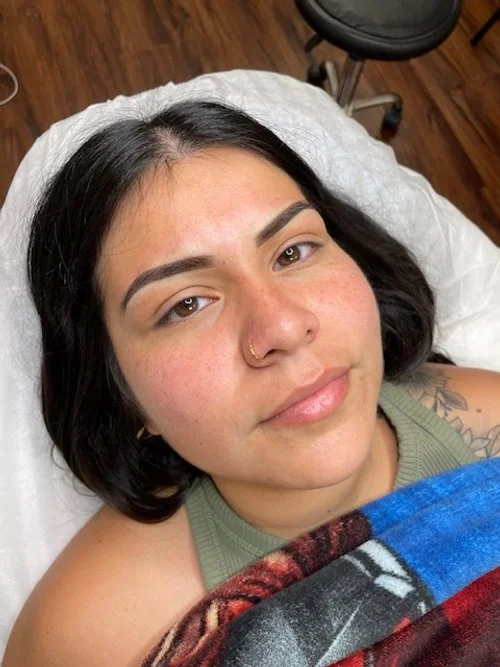 Close-up of a young woman lying on a medical bed with a white sheet, with dark hair, light skin, and a nose piercing, looking at the camera, with a blanket partly covering her chest.