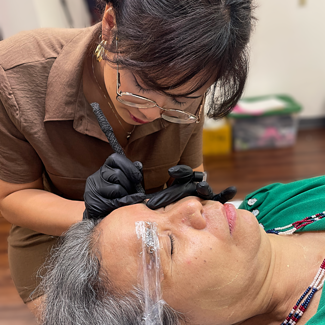 A woman getting inked on her face by a tattoo artist while lying down, with her eyes closed, wearing rings and black gloves, an apparent cosmetic tattoo procedure.