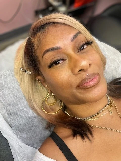 Close-up of a woman with blonde and brown hair, wearing gold jewelry, with makeup and a pierced lip, lying on a pillow.