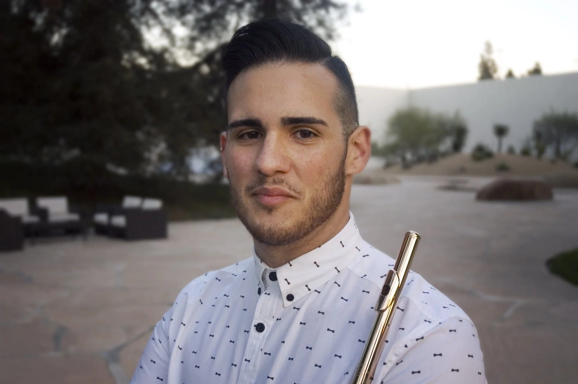 A young man with styled dark hair, light beard, and light skin, holding a gold-colored flute in an outdoor setting during sunset or late afternoon, with trees, rocks, and a white building in the background.
