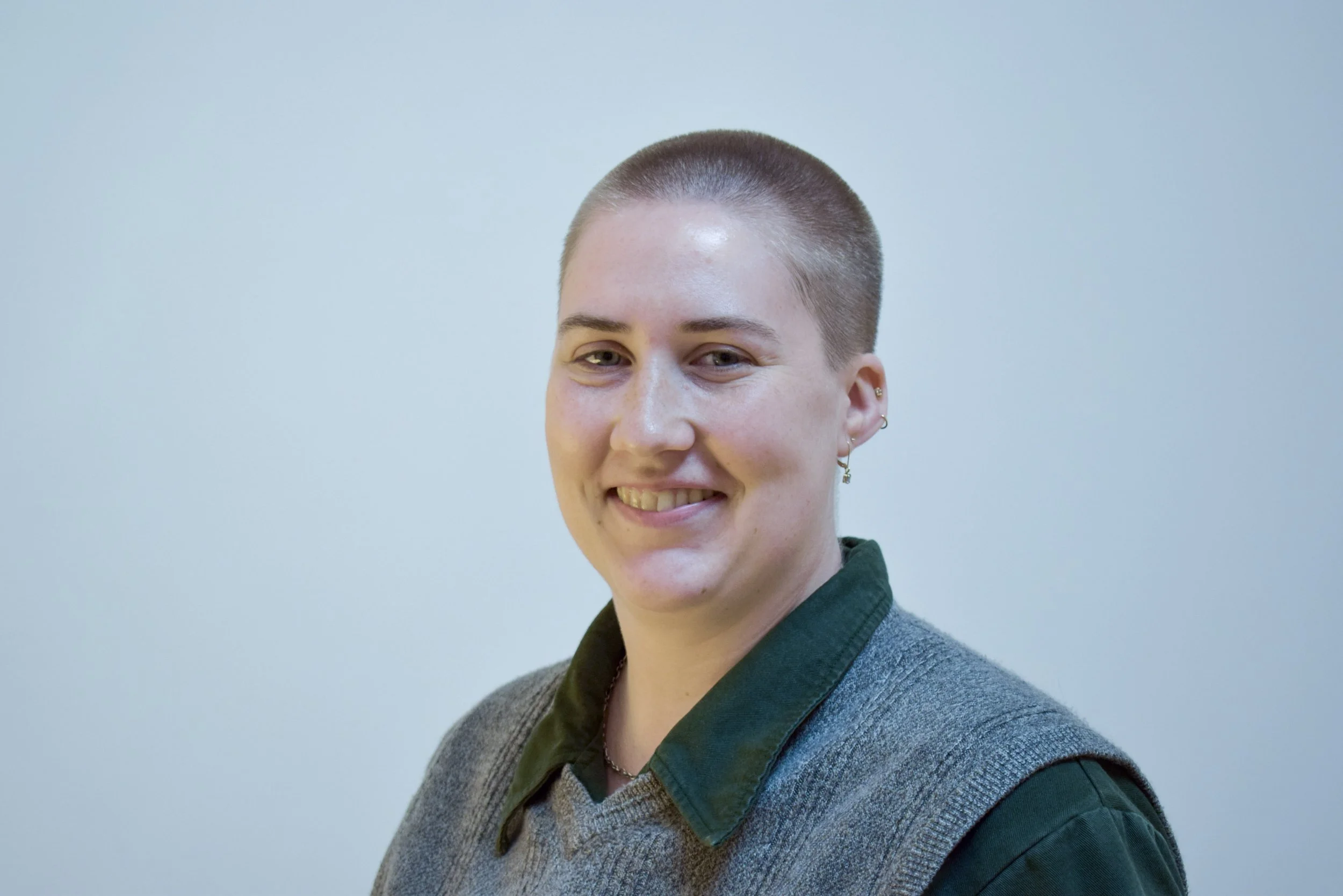 LGBTQIA-affirming therapist specializing in working with couples in Chicago  A person with a short buzz haircut smiling and looking at the camera, wearing a green collared shirt and a gray blazer, against a light blue background.