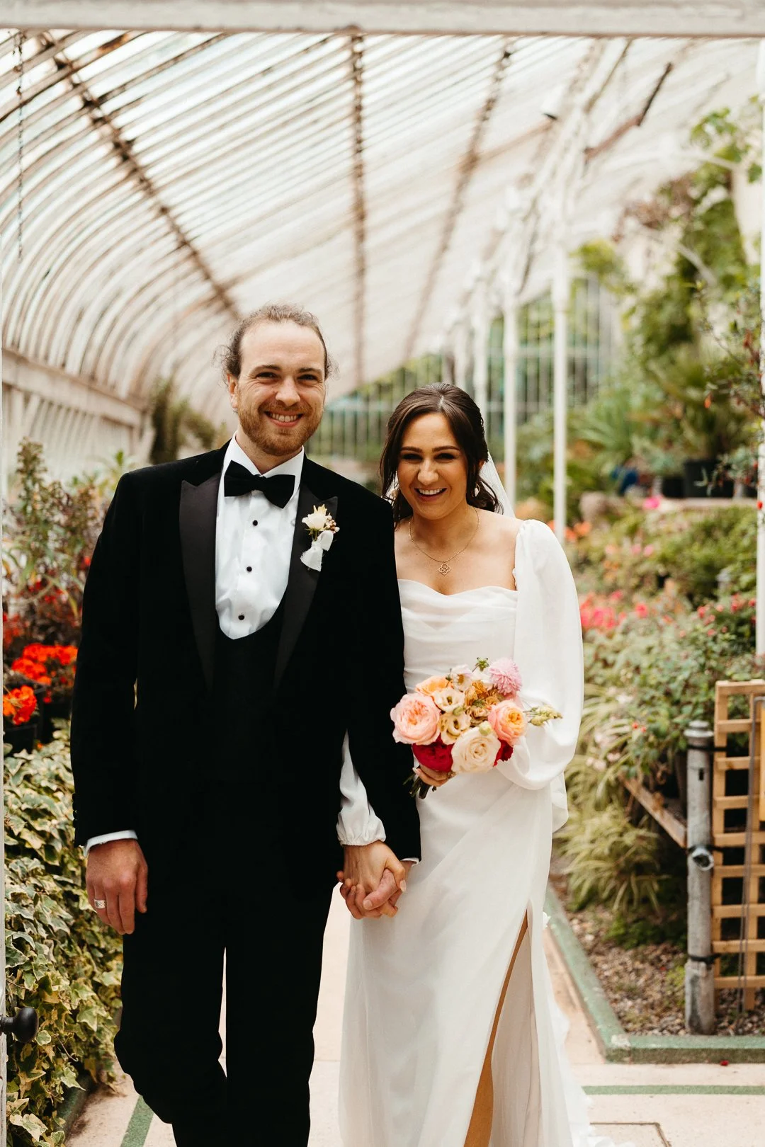 A newlywed couple walking hand in hand in a greenhouse filled with flowers, smiling happily.