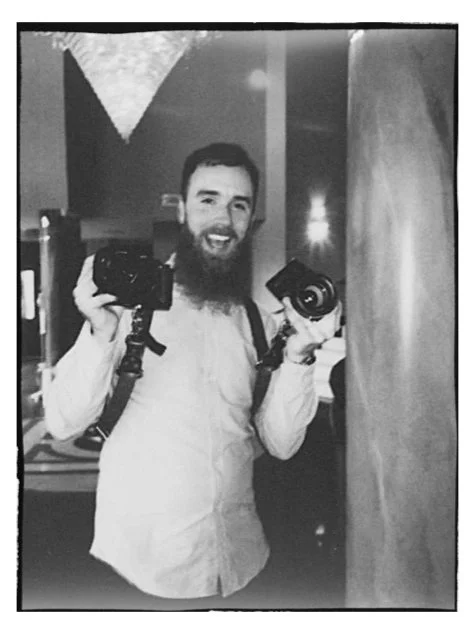 A man with a beard holding two professional cameras, smiling in a dimly lit indoor setting.