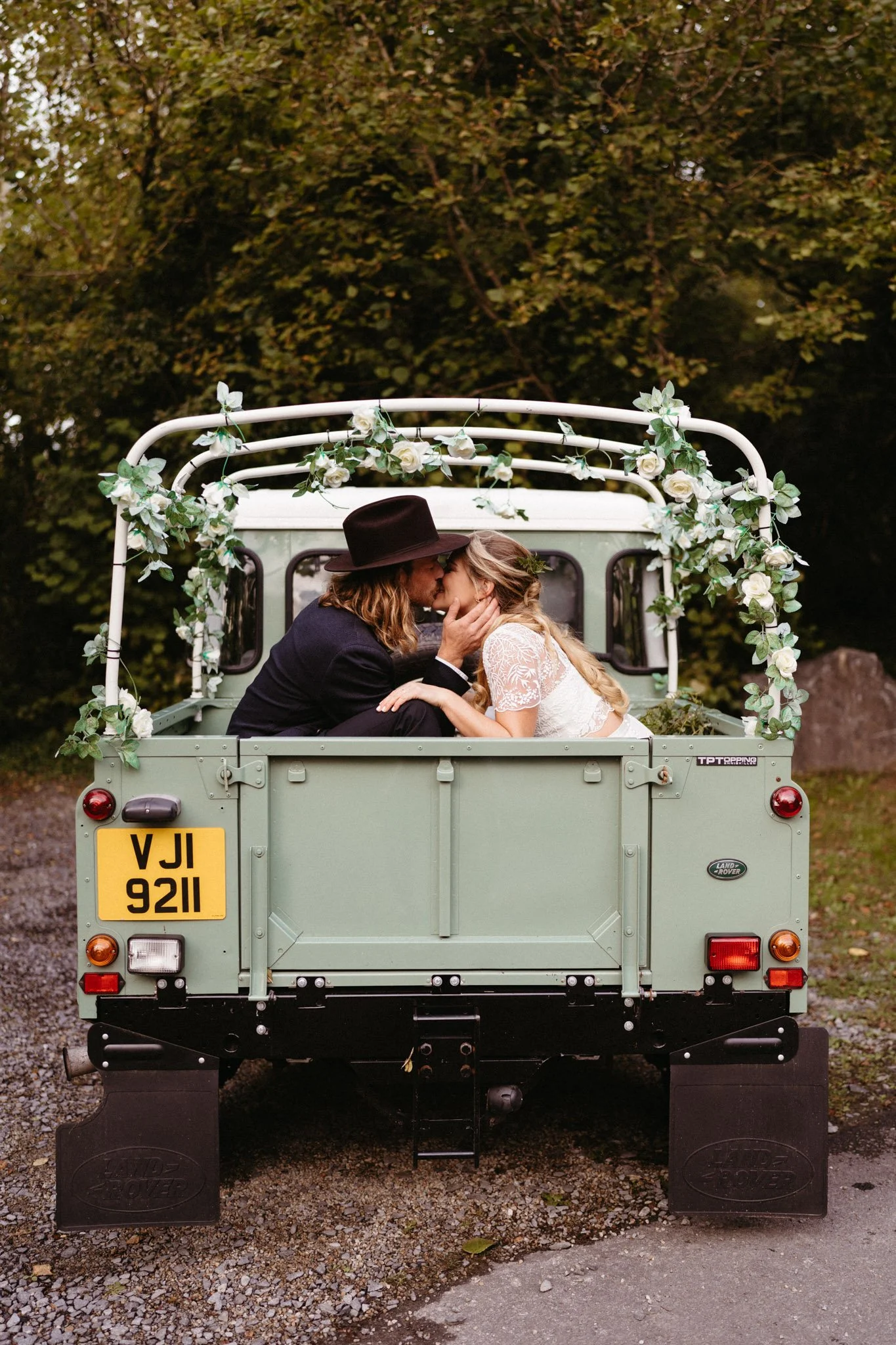 A couple in wedding attire sharing a kiss in the back of a vintage Land Rover decorated with white roses and greenery, outdoors in a wooded area.