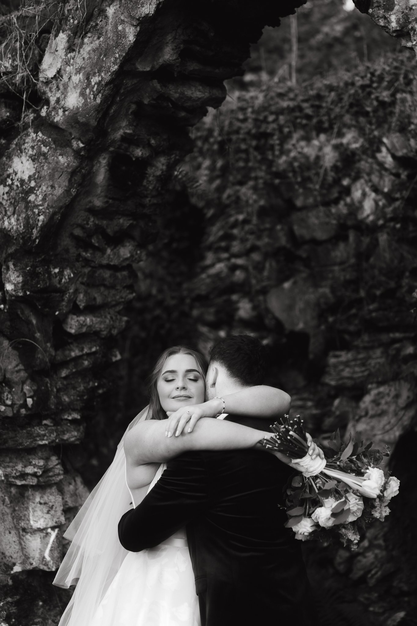 A bride and groom embrace, with the bride holding a bouquet of flowers, in an outdoor setting with rocky formations.