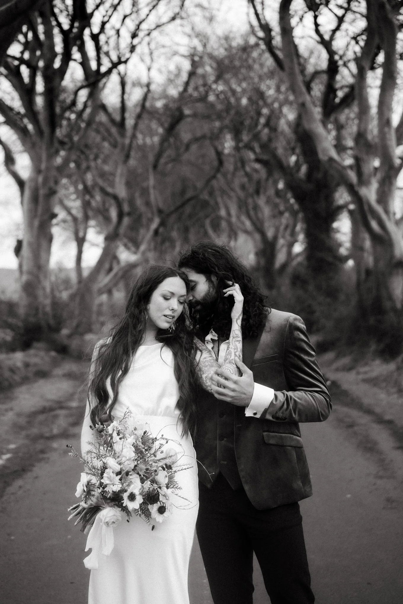 A black-and-white photo of a couple on their wedding day standing on a wooded path. The woman in a white dress holds a bouquet of flowers, and the man in a dark suit gently touches her face and chest as they lean their foreheads together.