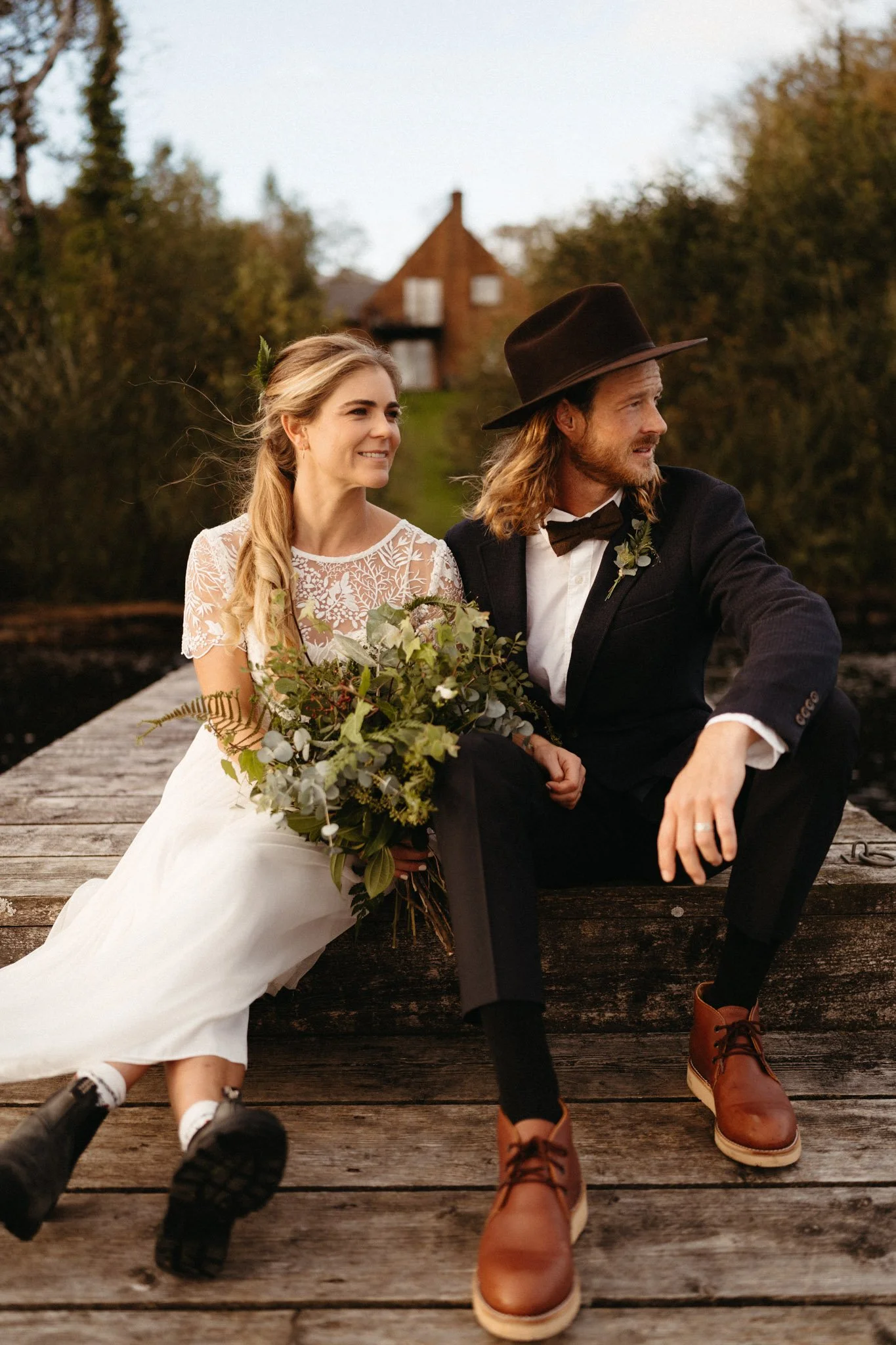 A couple dressed in wedding attire sitting on a wooden dock with greenery and a house in the background, enjoying a moment together.
