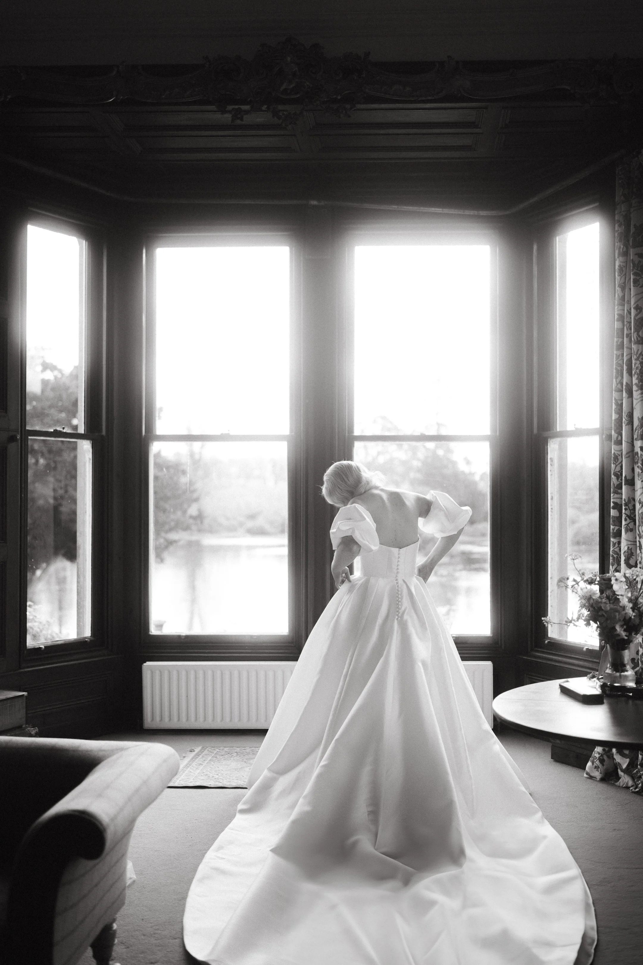 A woman in a wedding dress standing by large windows in a room with dark wood paneling, natural light streaming in.