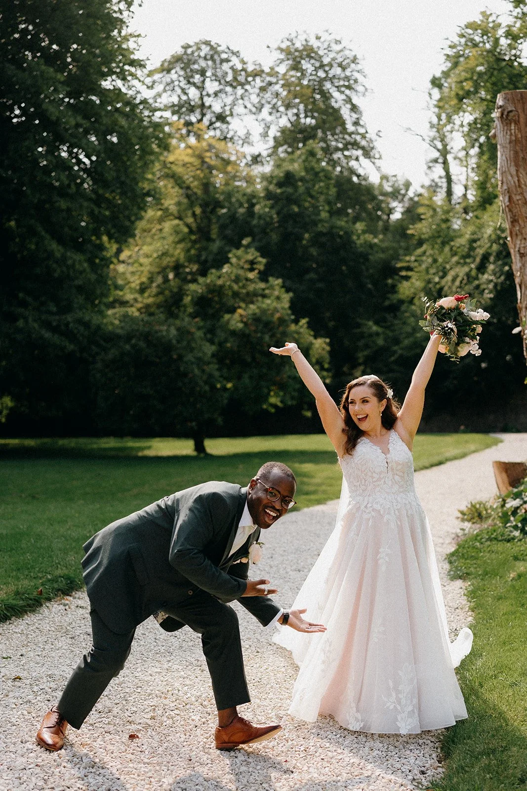 A bride in a white wedding dress with her arms raised, holding a bouquet of flowers, smiling joyfully. A man in a dark suit is crouching near her, pointing towards her with a big smile. They are outdoors on a gravel path surrounded by green grass and