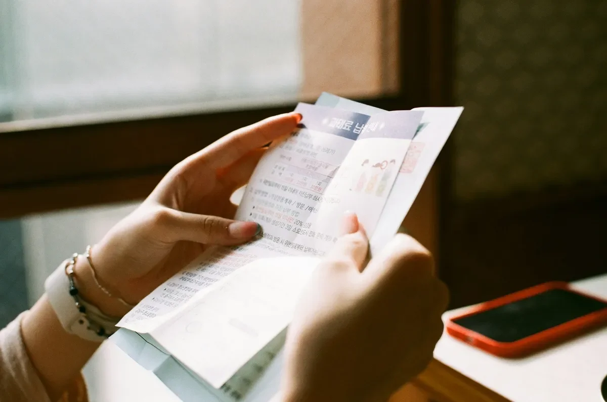 A person holding a crumpled paper or receipt with both hands, sitting at a table near a window, with a red smartphone on the table.