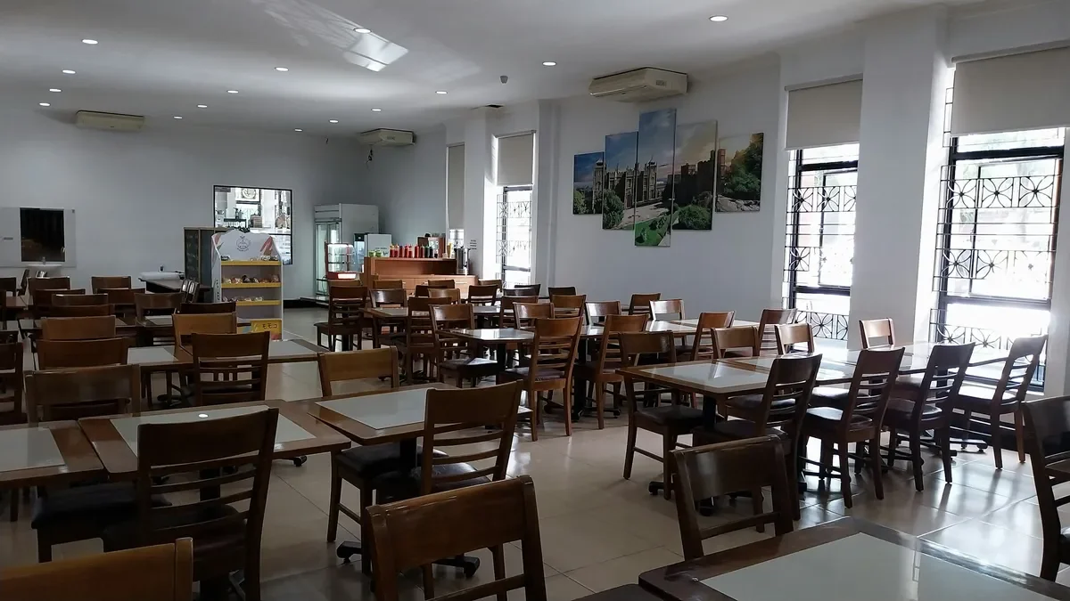 Empty restaurant with wooden tables and chairs, large windows with blinds, landscape art on the wall, and a dessert display counter.
