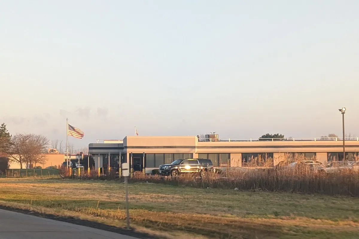 A single-story building with an American flag flying in front, cars parked outside, and a clear sky in the background.