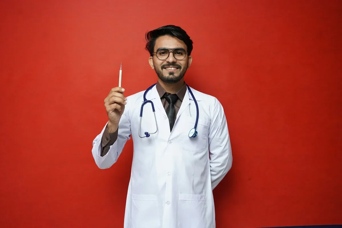 A male doctor with glasses, a beard, and a mustache, wearing a white coat and a stethoscope around his neck, holding a syringe in his right hand, standing against a red background, smiling.