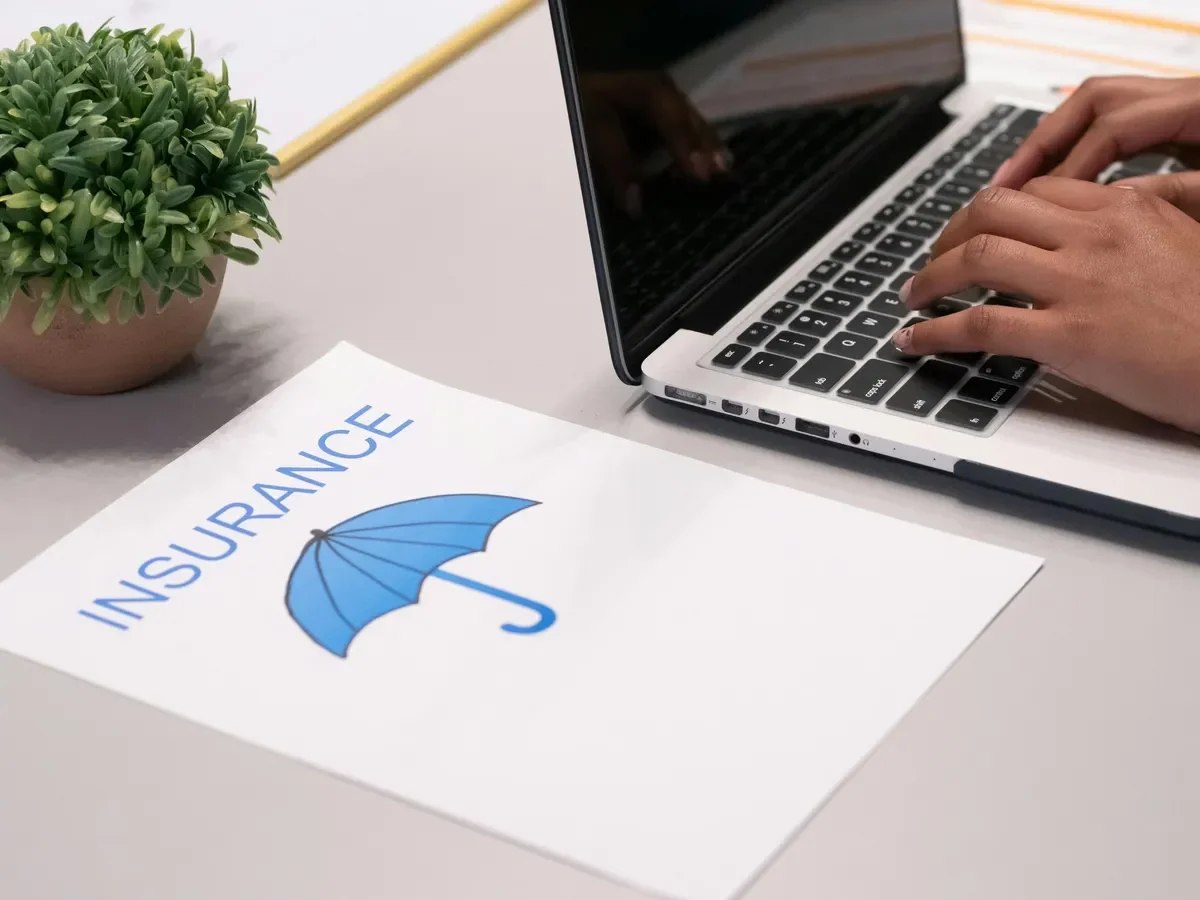 A clipboard with insurance paperwork featuring a blue umbrella graphic, a potted plant, and a person's hands typing on a laptop keyboard on a desk.