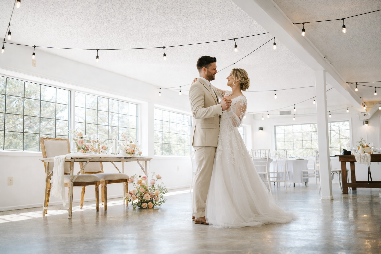 A bride and groom dance together in a bright, decorated wedding venue with large windows, string lights, and floral arrangements.