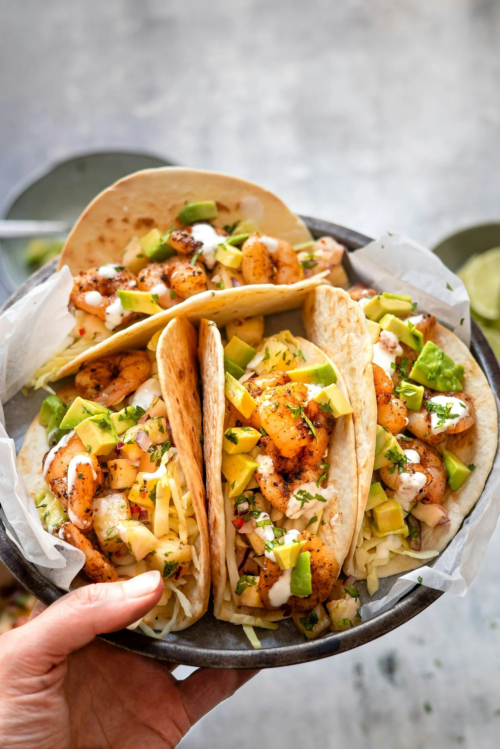 A person holding a basket with three shrimp tacos topped with diced avocado, chopped cilantro, and a drizzle of sauce.