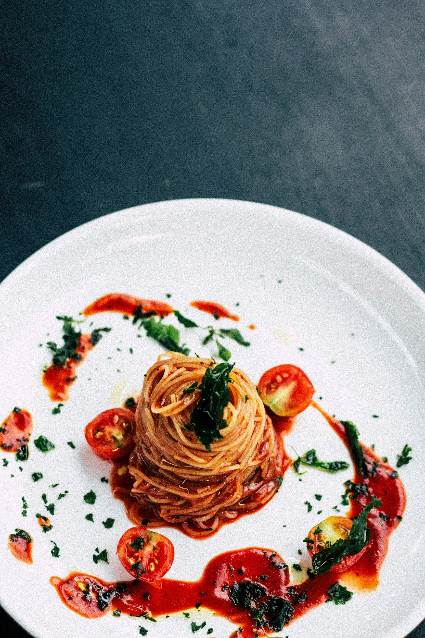 A white plate with a small mound of spaghetti pasta topped with basil, garnished with cherry tomatoes, and drizzled with tomato sauce and chopped herbs.