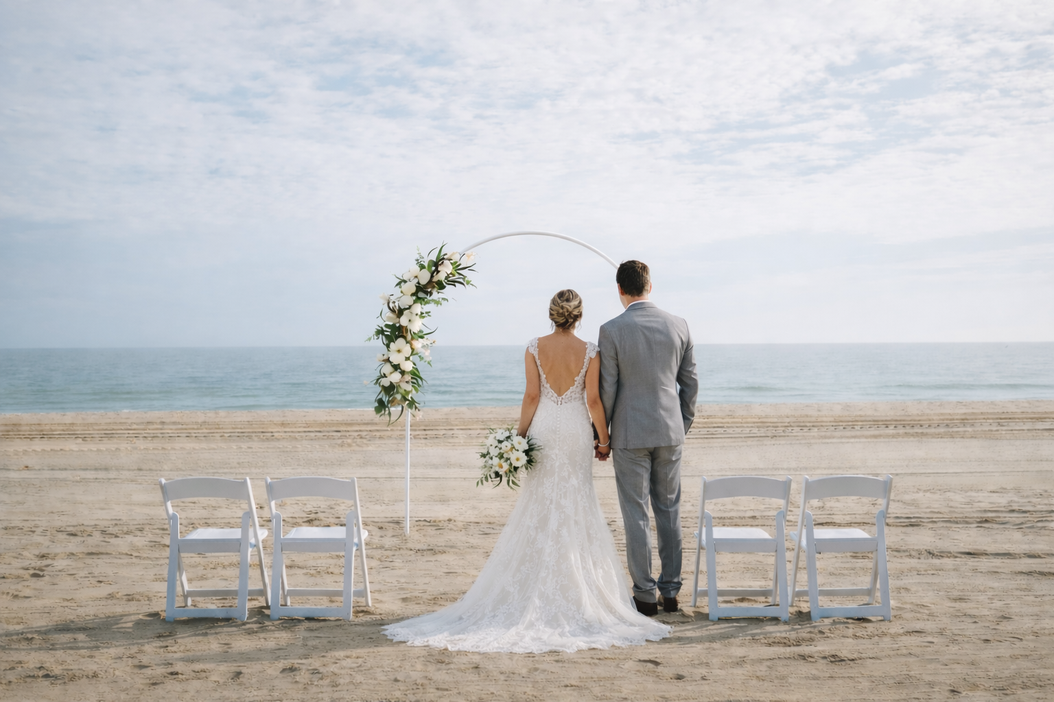 A bride and groom holding hands on a sandy beach, facing the ocean, during their wedding ceremony. They are standing under a white floral arch with white chairs on either side.