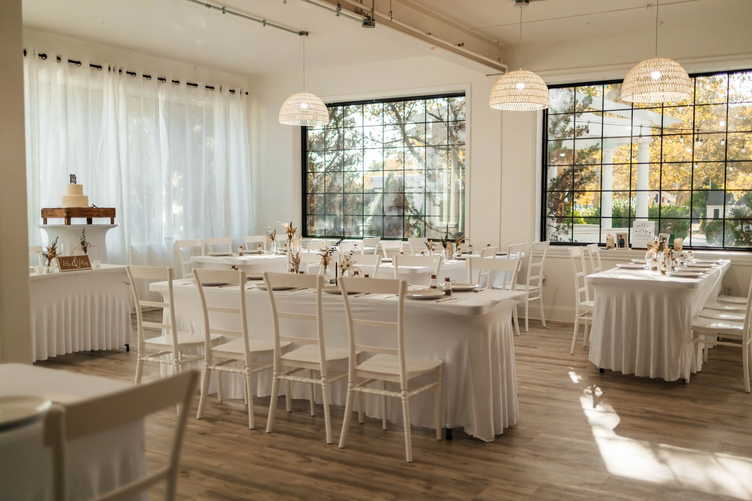 Elegant indoor wedding reception setup with white tables and chairs, floral centerpieces, a tiered wedding cake on a cake stand, and large windows with natural light.