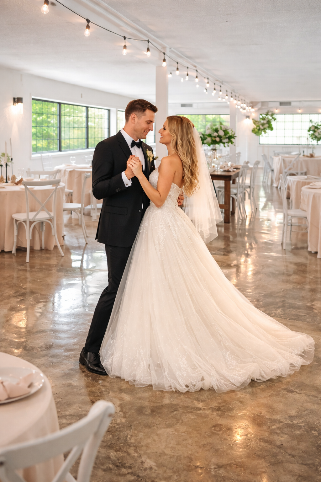 A bride and groom dancing together in a decorated wedding reception hall with tables, chairs, and string lights.
