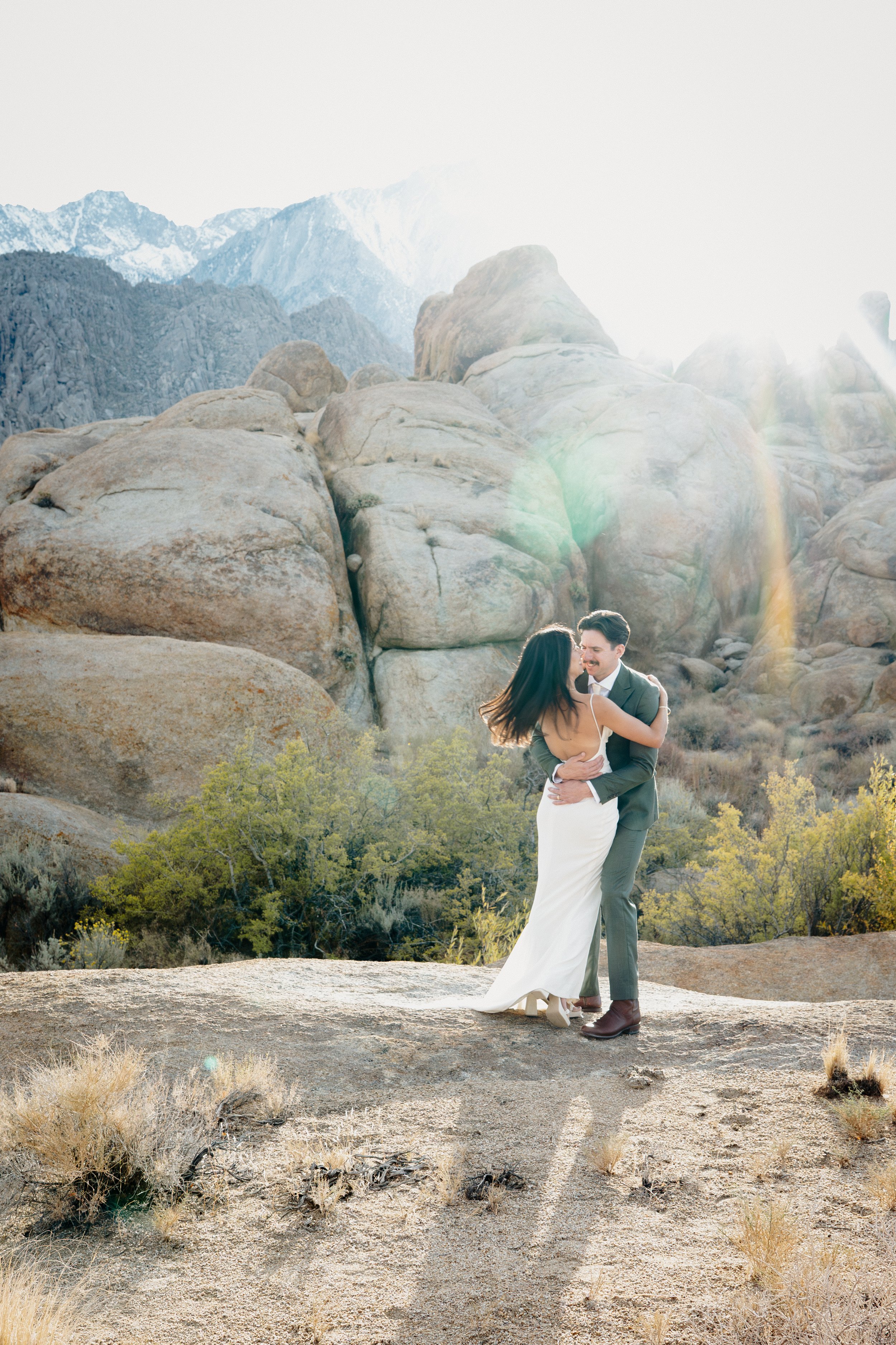A couple dancing outdoors in a mountainous desert landscape with large rocks and sparse vegetation, illuminated by bright sunlight.