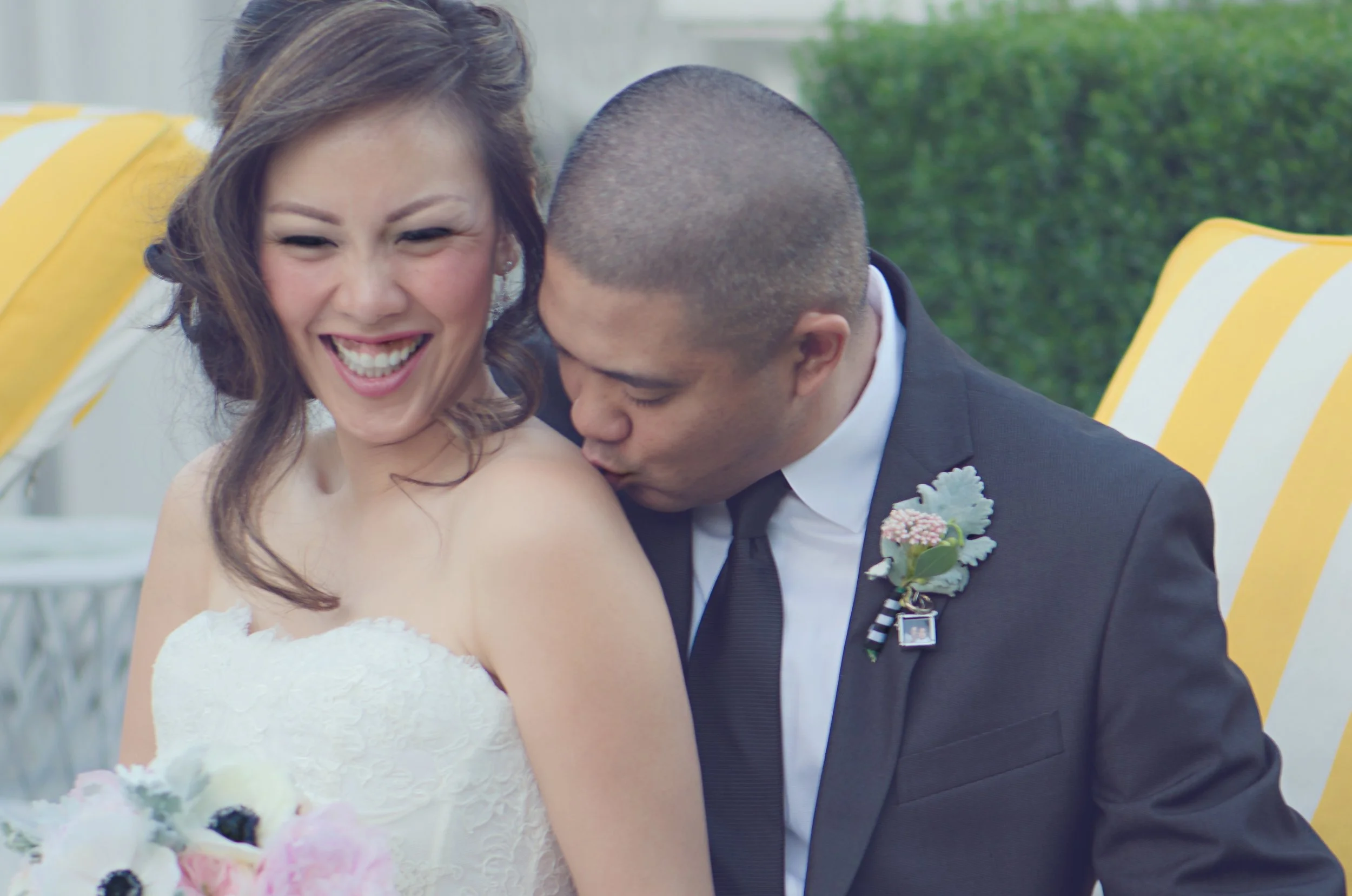 A bride and groom sharing a joyful moment during their wedding, with the groom kissing the bride's shoulder while she smiles and laughs.
