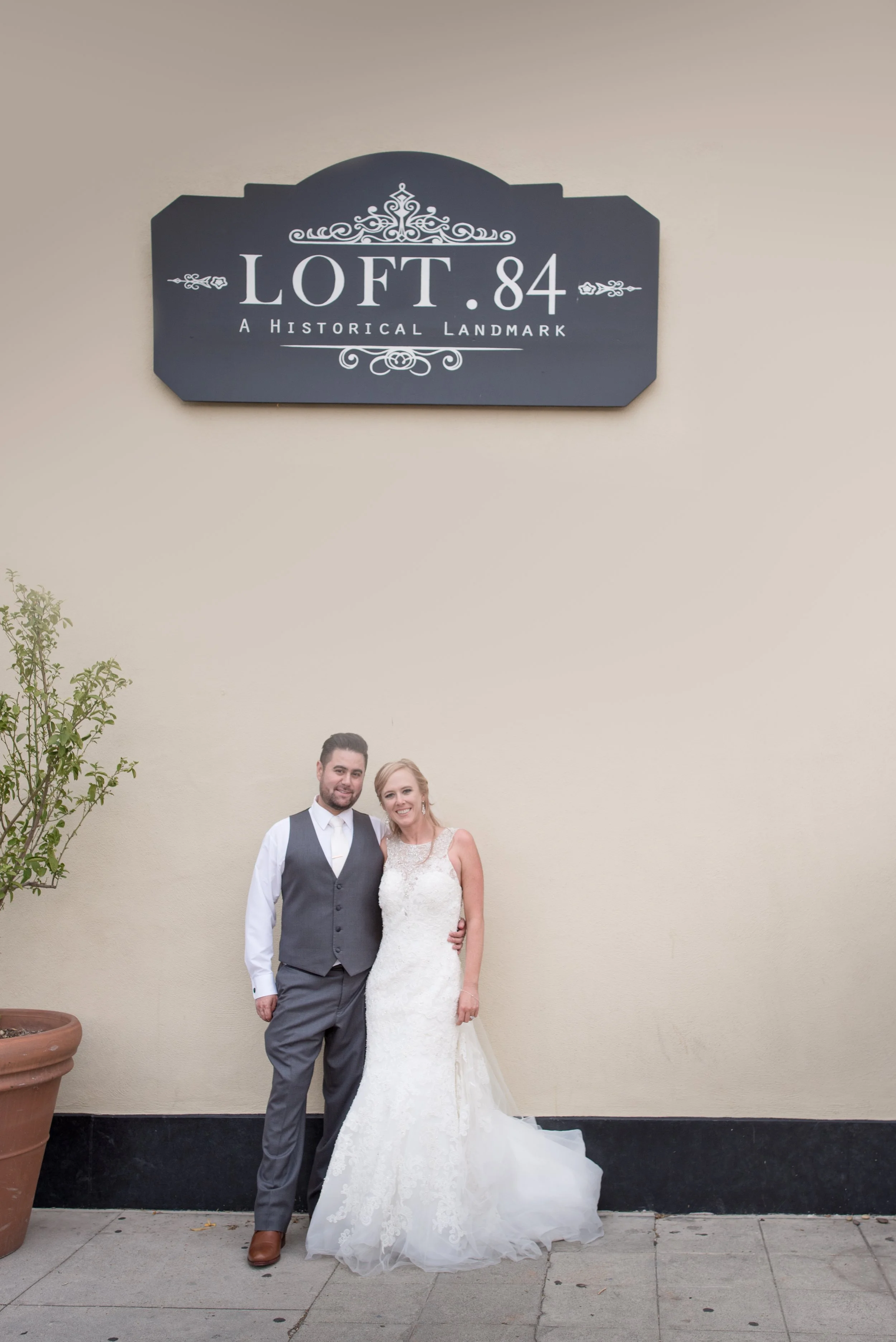 A bride and groom standing outside a building with a sign that reads 'LOFT.84 A Historical Landmark.' The bride is in a white lace wedding dress, and the groom is in a gray suit with a vest and tie. They are smiling and embracing each other.