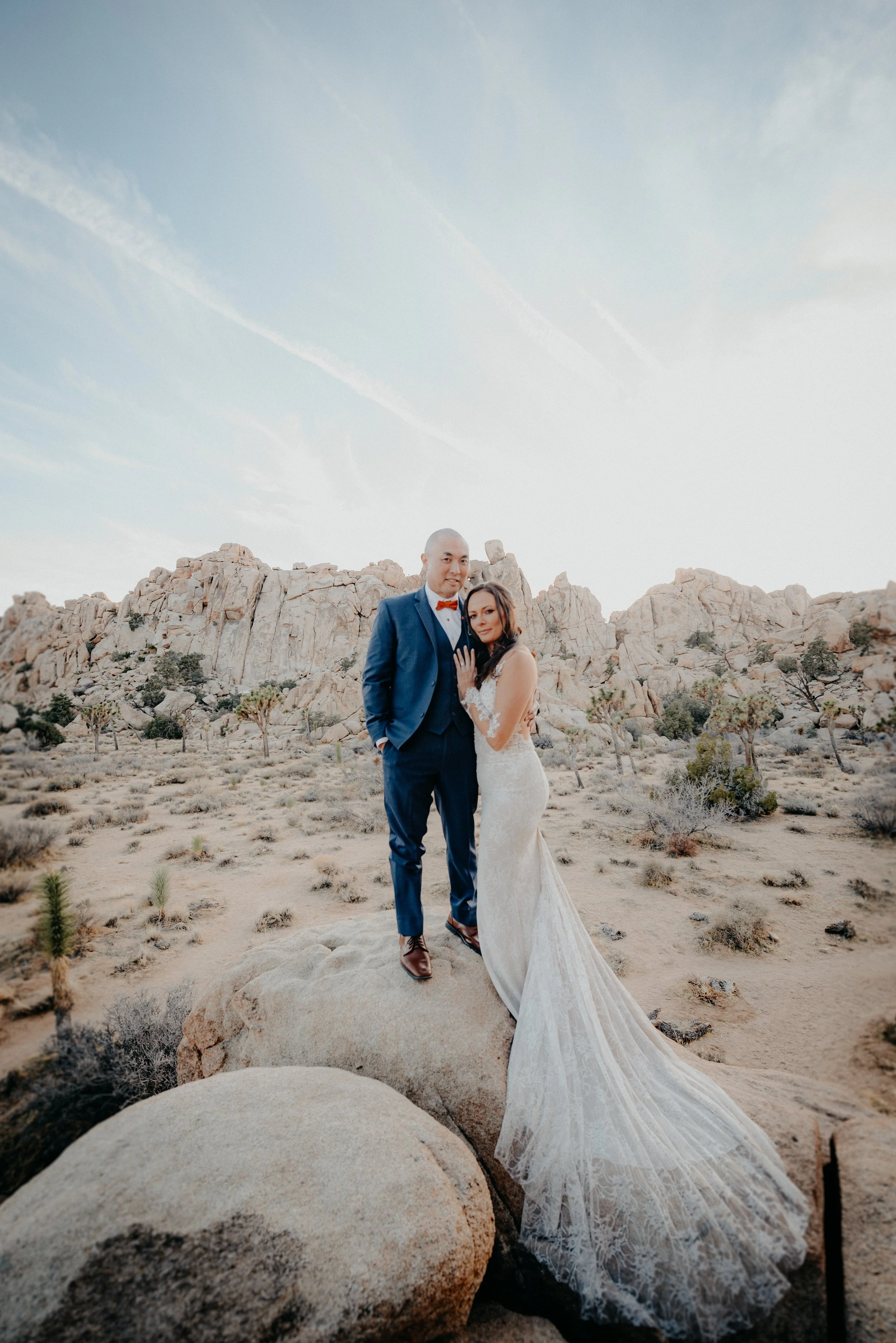 Brid couple standing on a large rock in a desert landscape with rocky hills and sparse vegetation under a blue sky with wispy clouds.