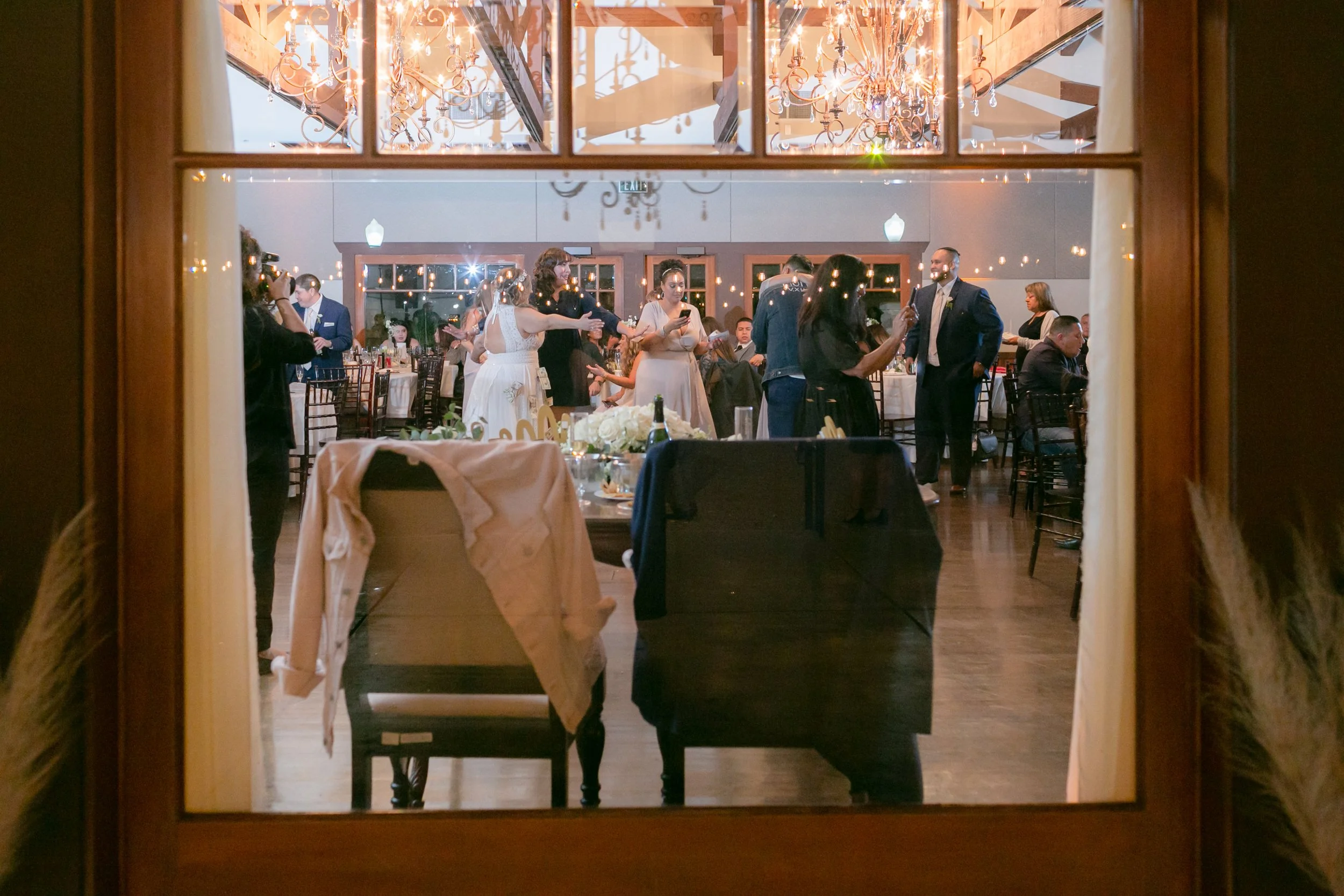 Guests dancing and socializing at a wedding reception seen through a window with curtains.