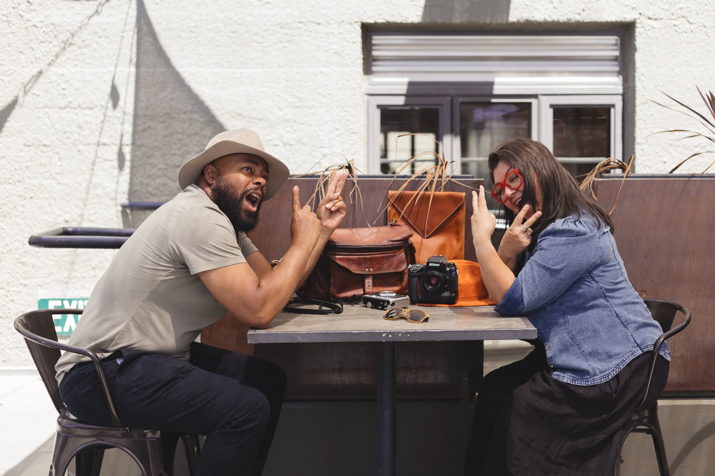 Two people sitting at a table outdoors, making peace signs. They have cameras and bags on the table. The man is wearing a beige hat and shirt; the woman is wearing red glasses and a denim jacket.