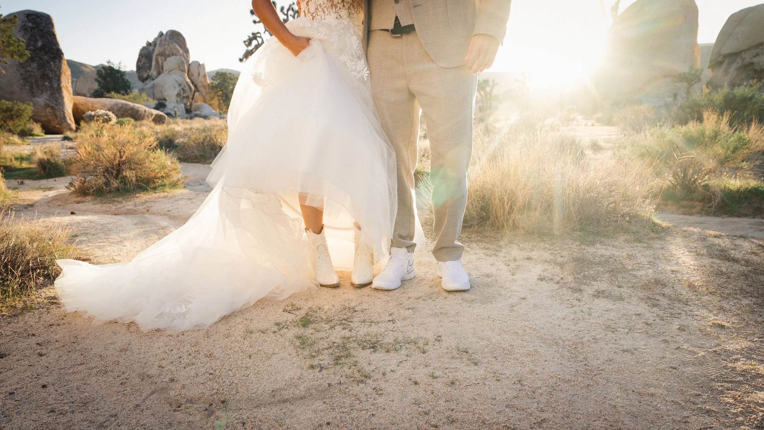 Bride and groom standing close together outdoors, with the bride lifting her wedding dress slightly, sunset lighting, rocky desert landscape, dry grass, and large rock formations in the background.