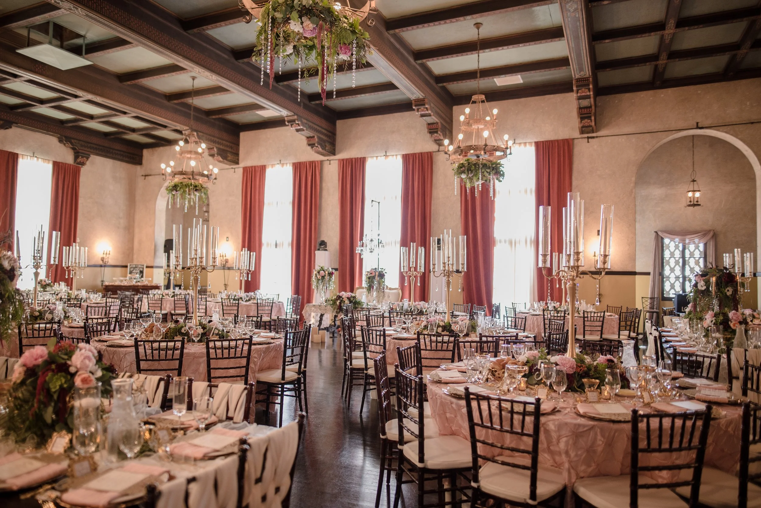 Elegant banquet hall decorated for event with round tables draped in pink tablecloths, detailed floral centerpieces, tall candelabras, and chairs with white cushions. The room features high ceilings with wooden beams, chandeliers, large windows with pink curtains, and hanging floral arrangements.