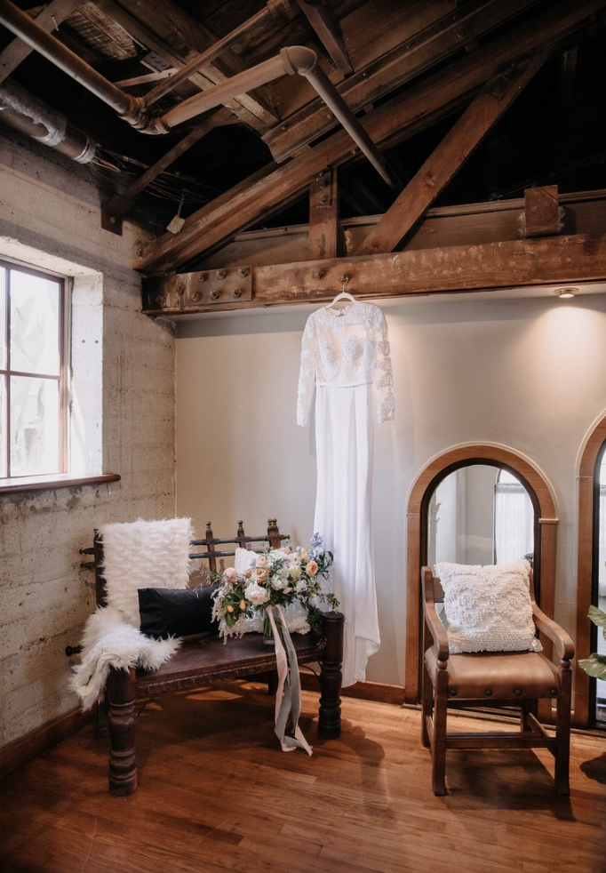 A white wedding dress hanging on a wall, with a bouquet of flowers on a wooden bench. The room has exposed wooden beams and a window with natural light, giving it a rustic feel.