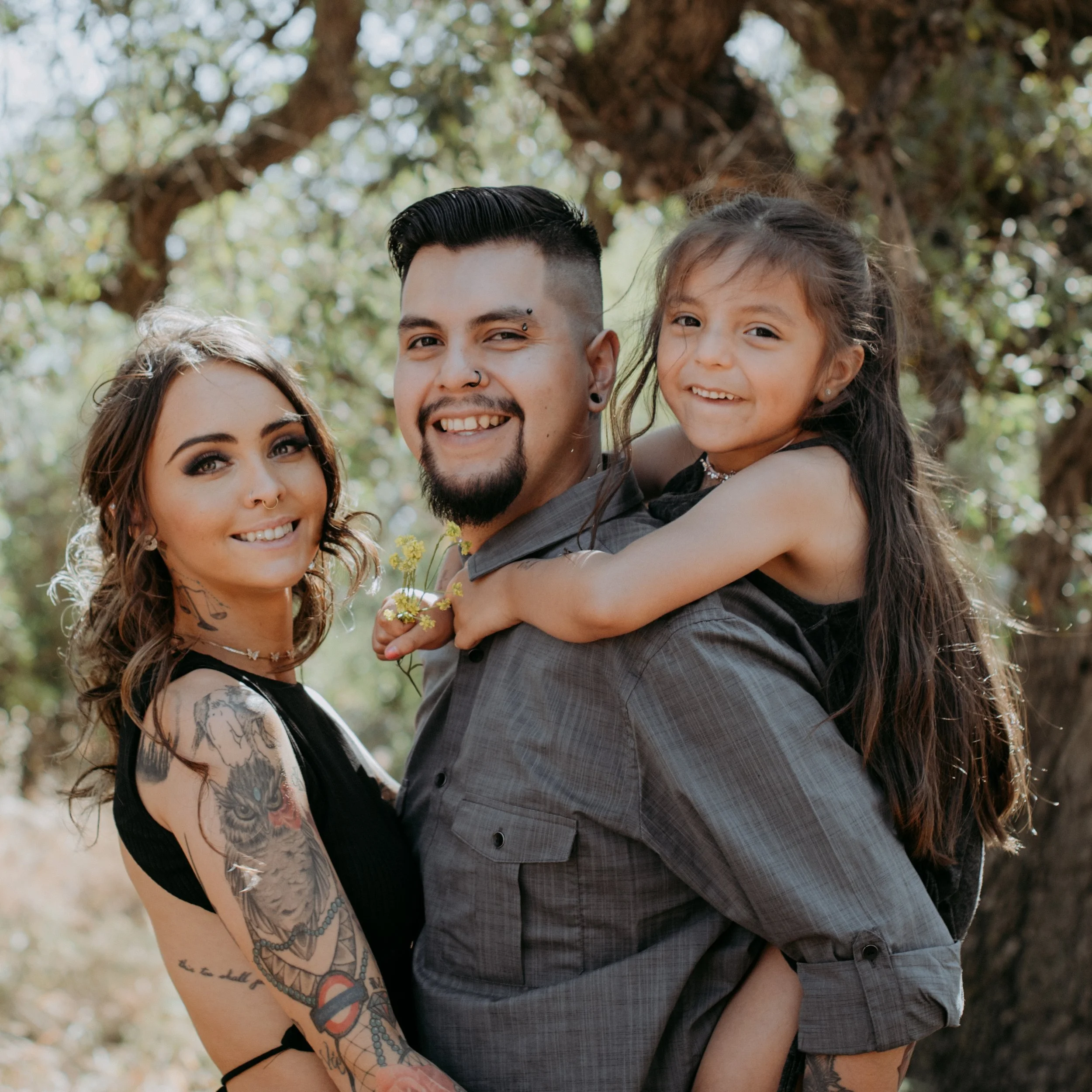 A diverse family of three smiling outdoors, with a woman with tattoos and a young girl on a man's back, all surrounded by trees.