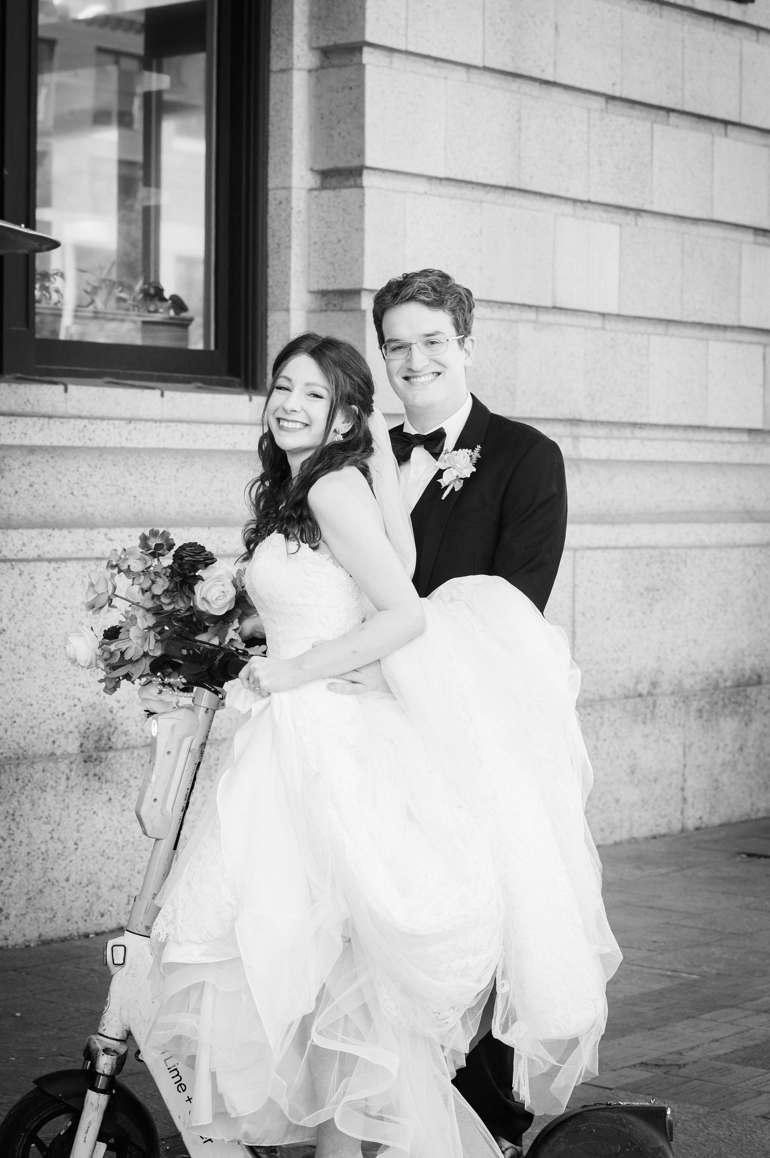 A newlywed couple, woman in a wedding dress and man in a tuxedo, smiling and posing outside a building, with the woman holding a bouquet of flowers.