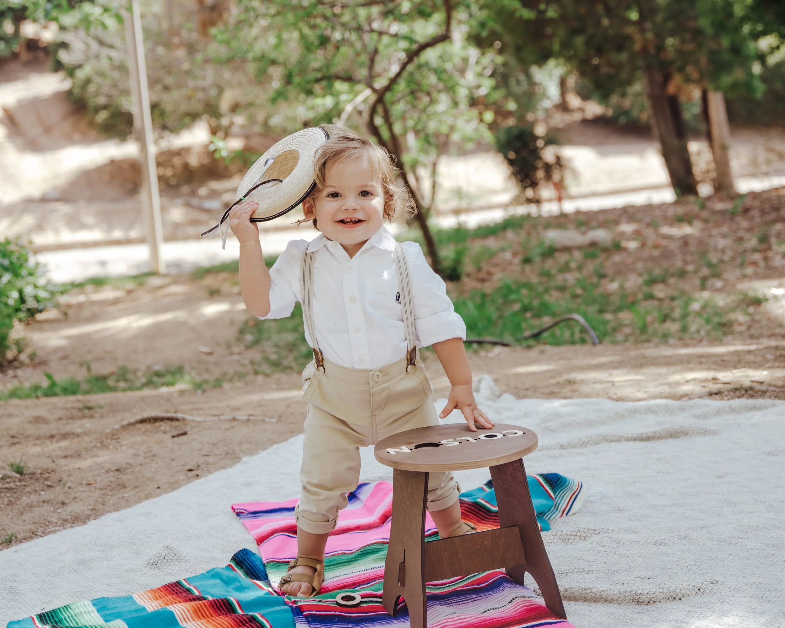 toddler taking his hat off standing by a stool on top of a mexican rug, and white blanket