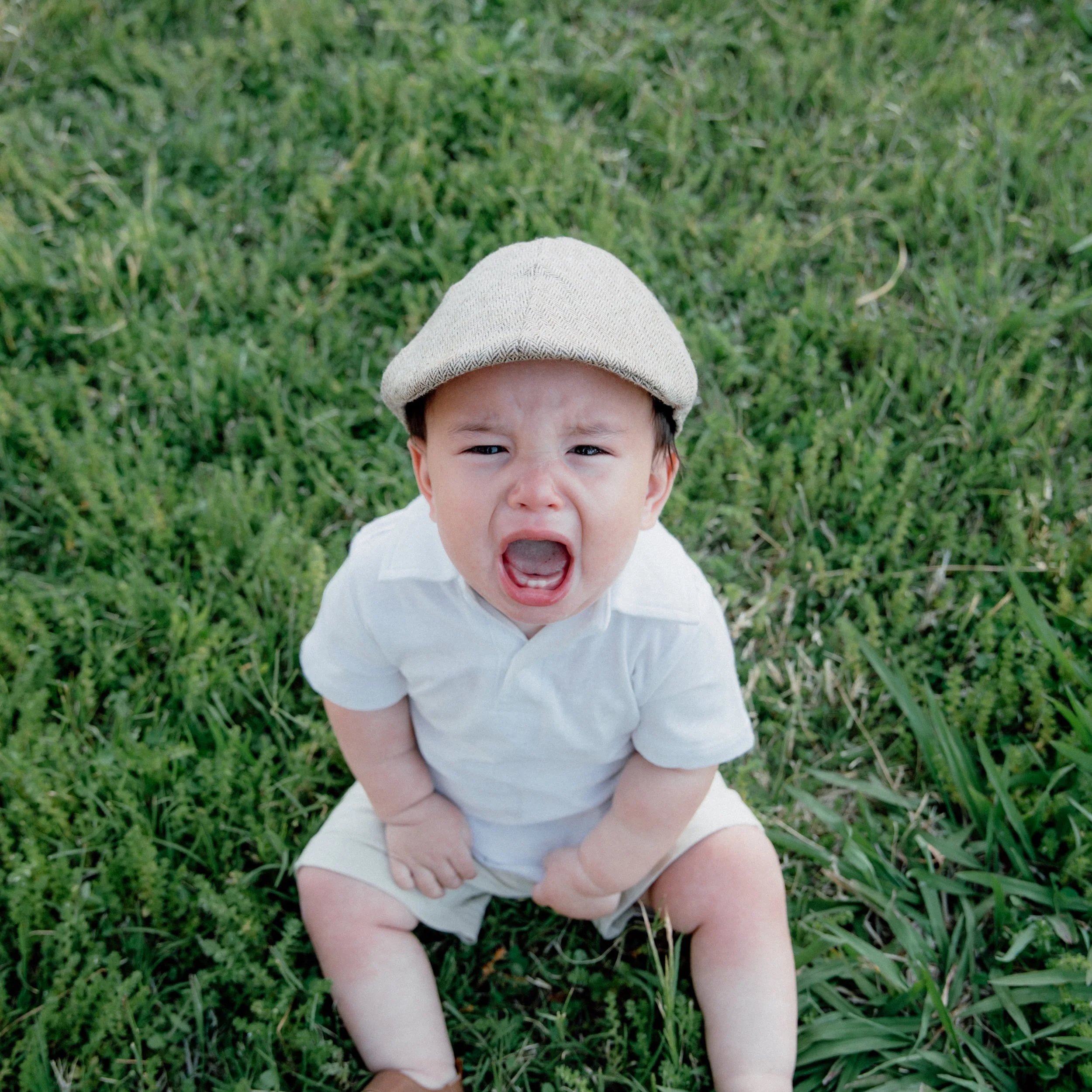 A young child with a beige cap and white shirt, sitting on green grass, appears to be crying or upset.