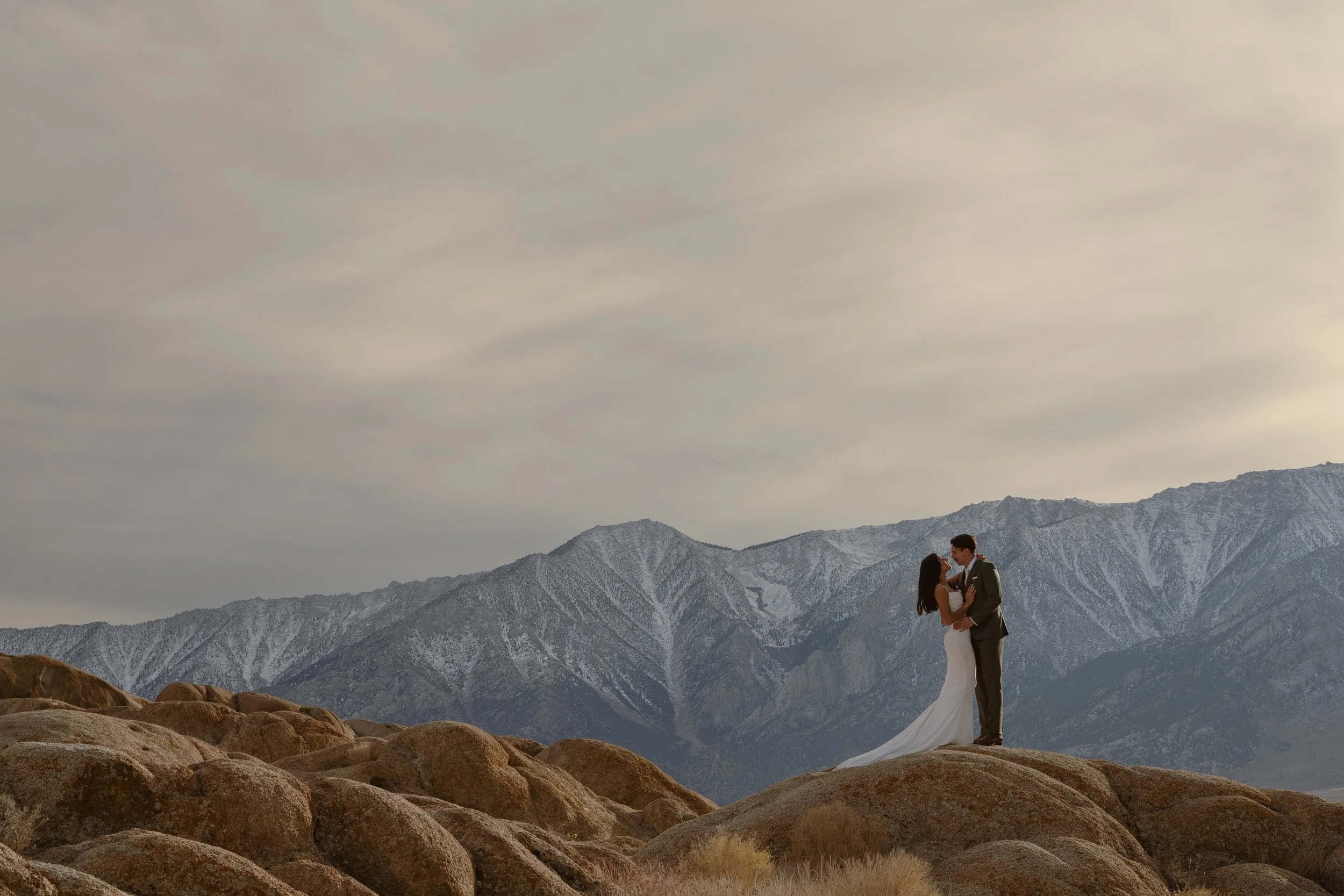 A bride and groom standing on rocks in a mountainous landscape, holding each other and gazing at each other, with snow-capped mountains in the background and a cloudy sky overhead.