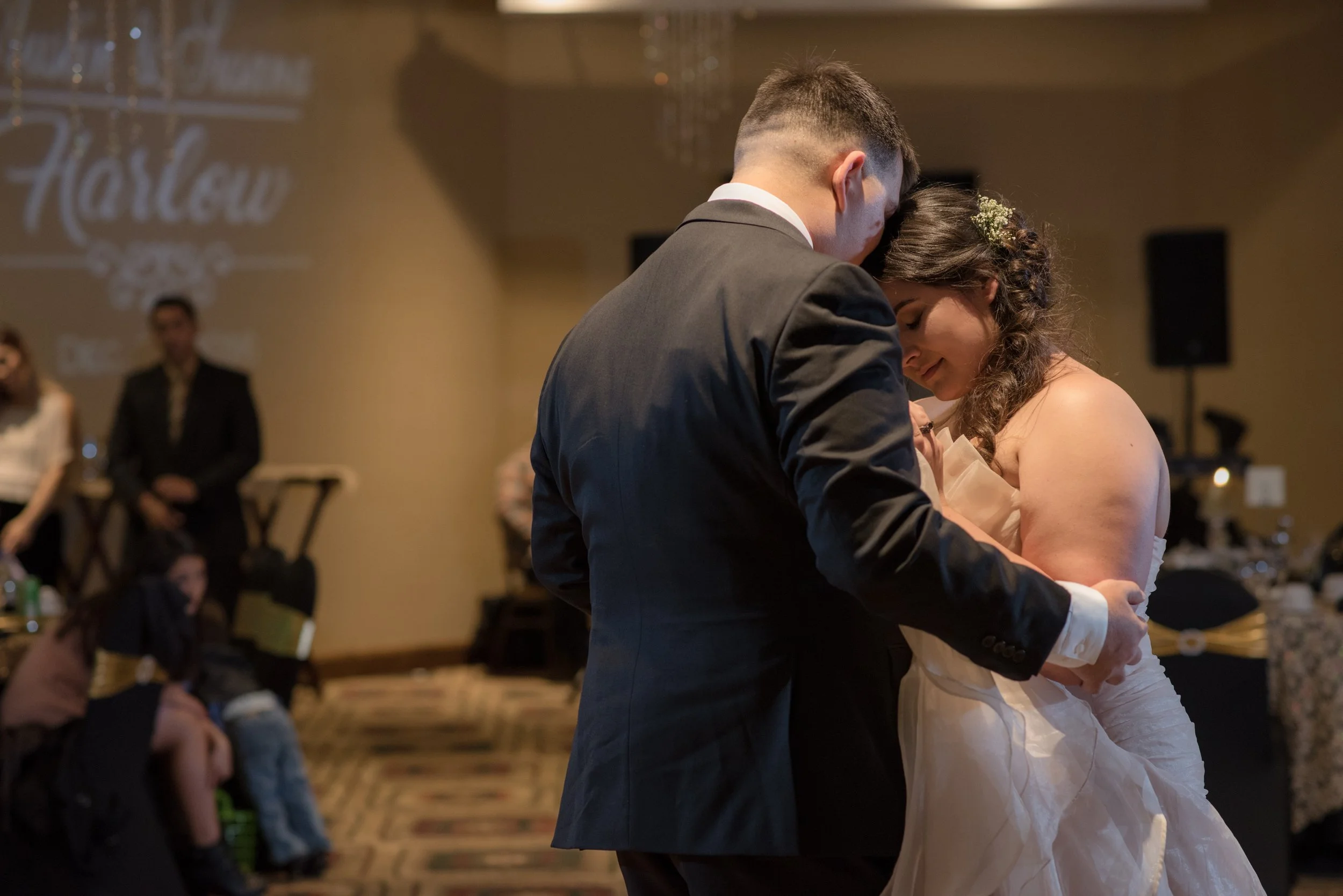 A bride and groom dance closely at their wedding reception, with guests watching in the background.