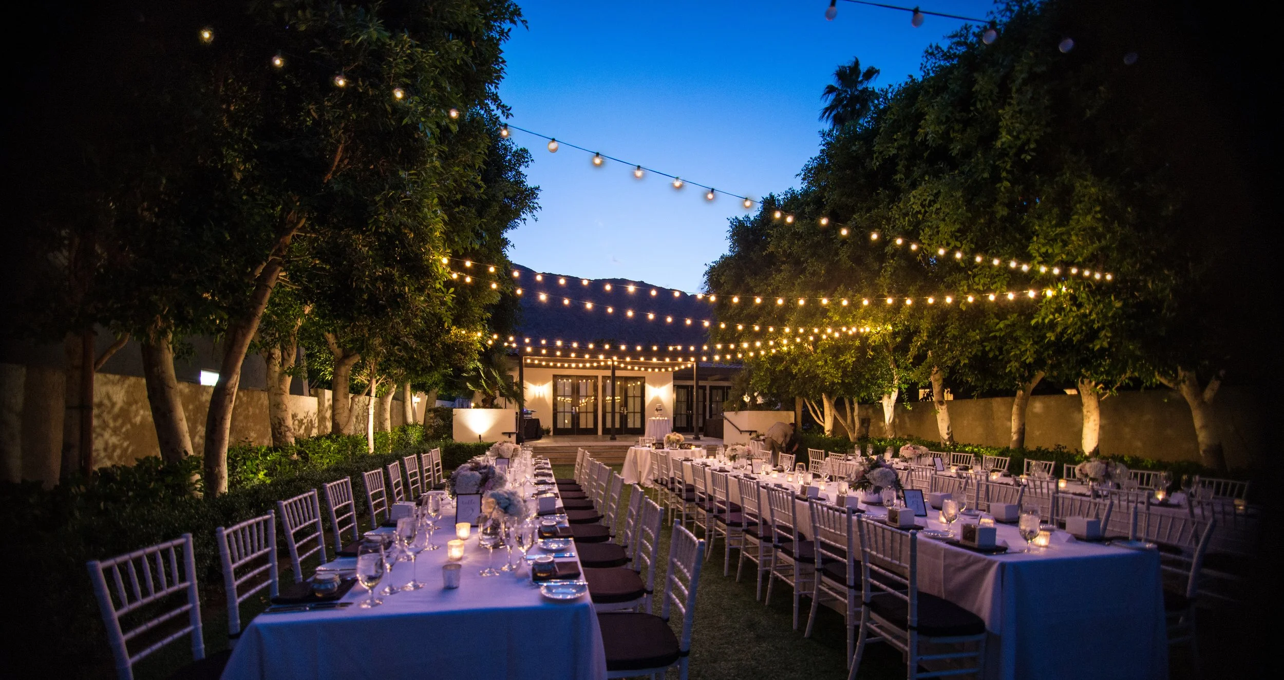 Outdoor evening event with long banquet tables set with white tablecloths, glassware, candles, and small floral arrangements, surrounded by trees with string lights overhead, leading to a building with glass doors.