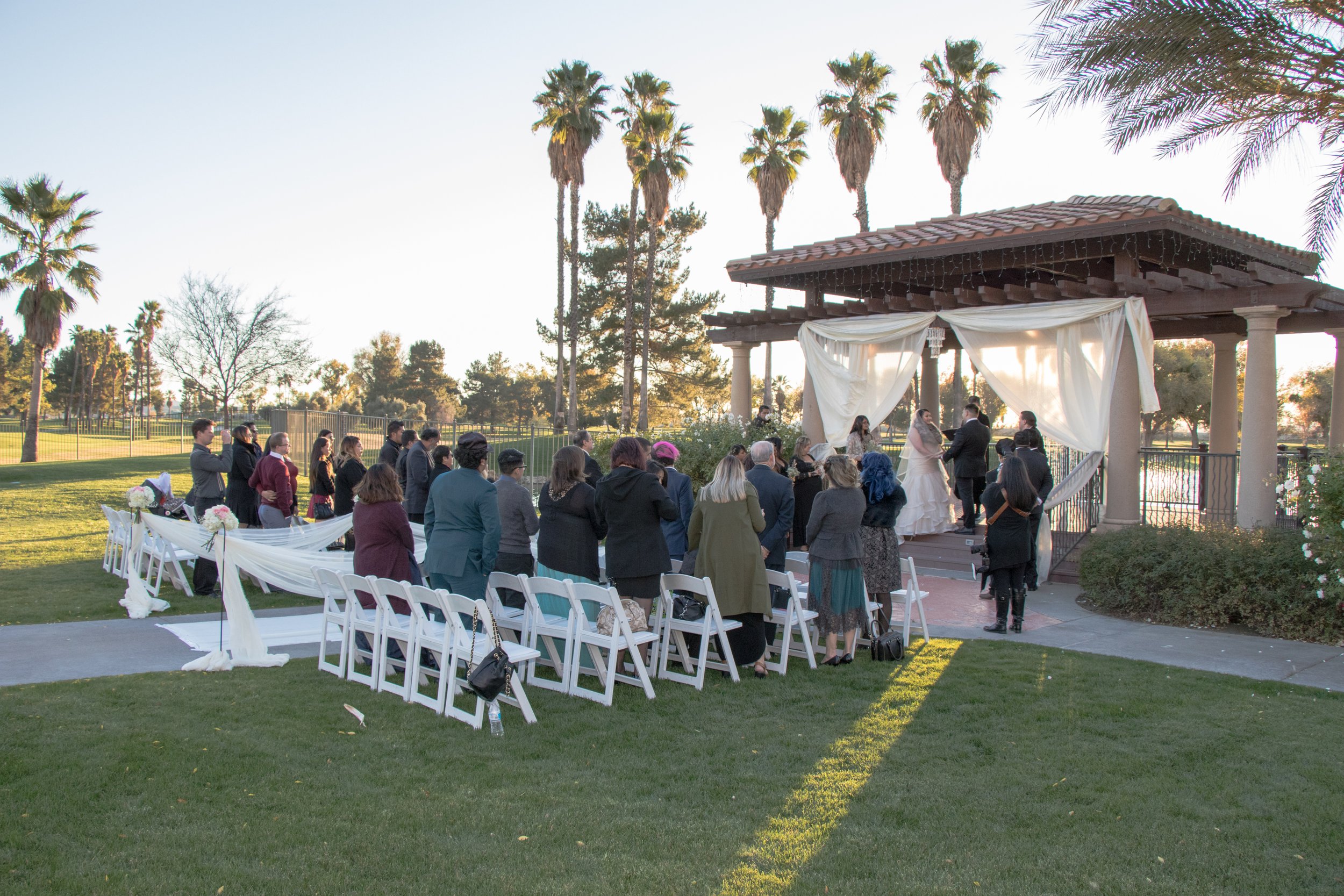 Outdoor wedding ceremony on a gazebo with white drapery, guests standing and seated on white chairs, palm trees and a lake in the background, during late afternoon.