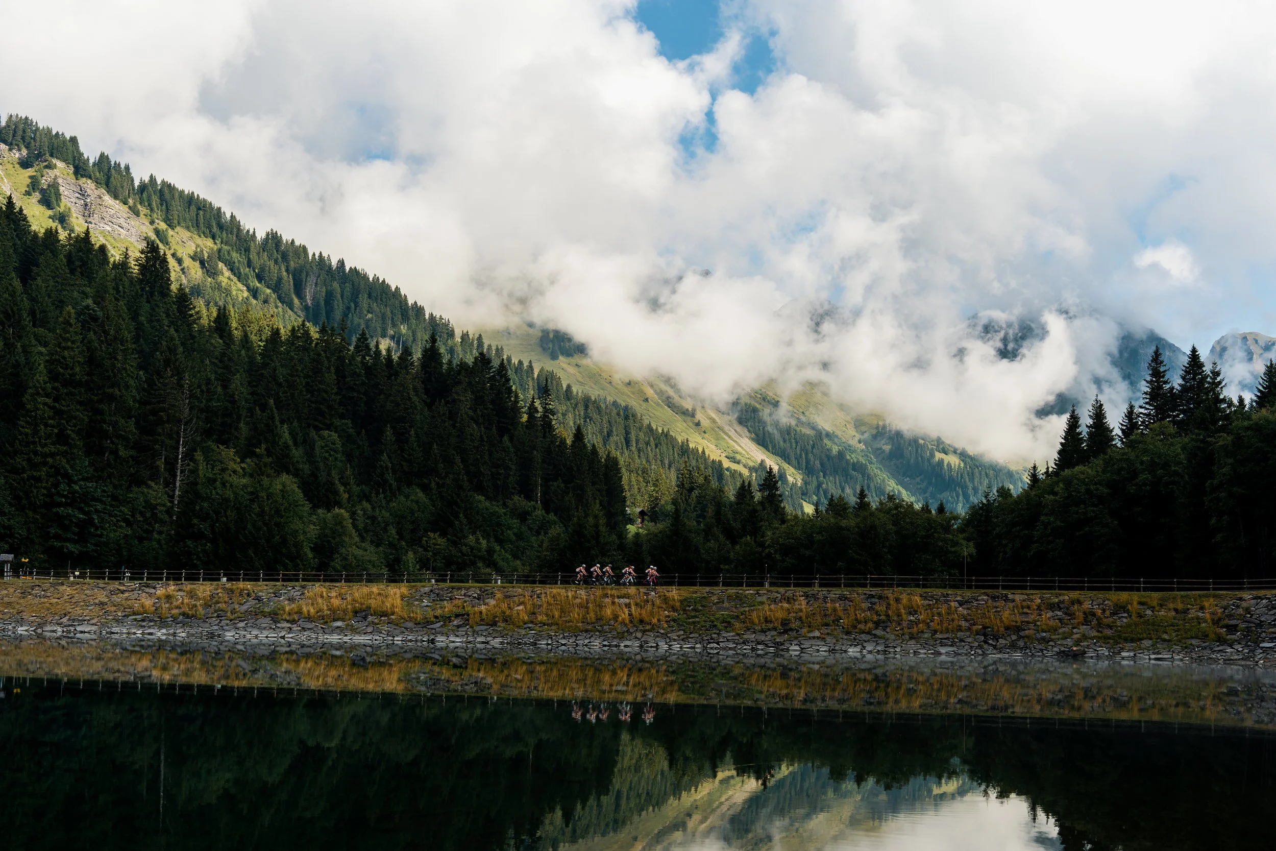 Mountain landscape with dense green trees, misty clouds, and a calm lake reflecting the scenery. There are cyclists riding along the road near the lake.