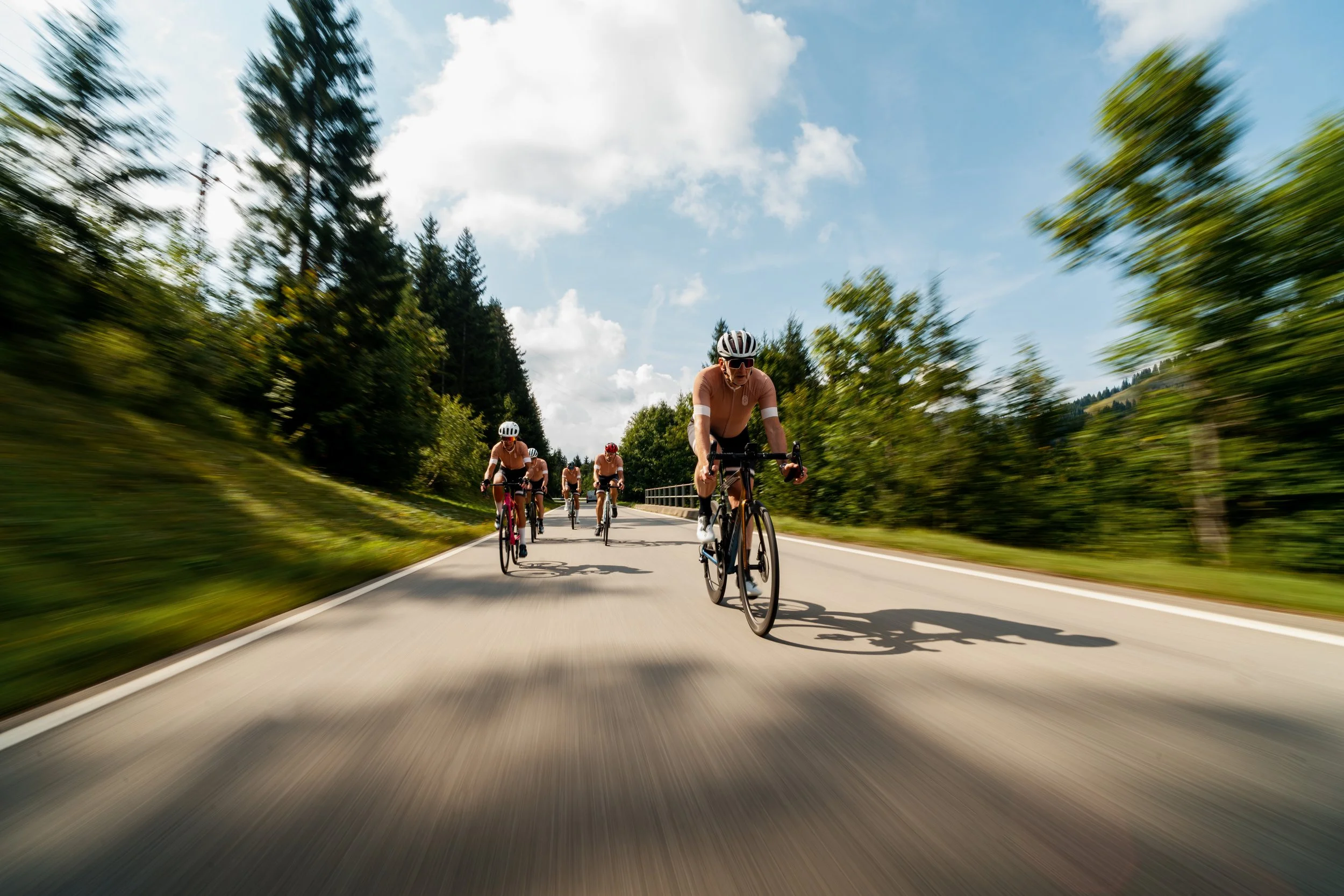 A group of cyclists riding on a winding road surrounded by lush trees and a partly cloudy sky.