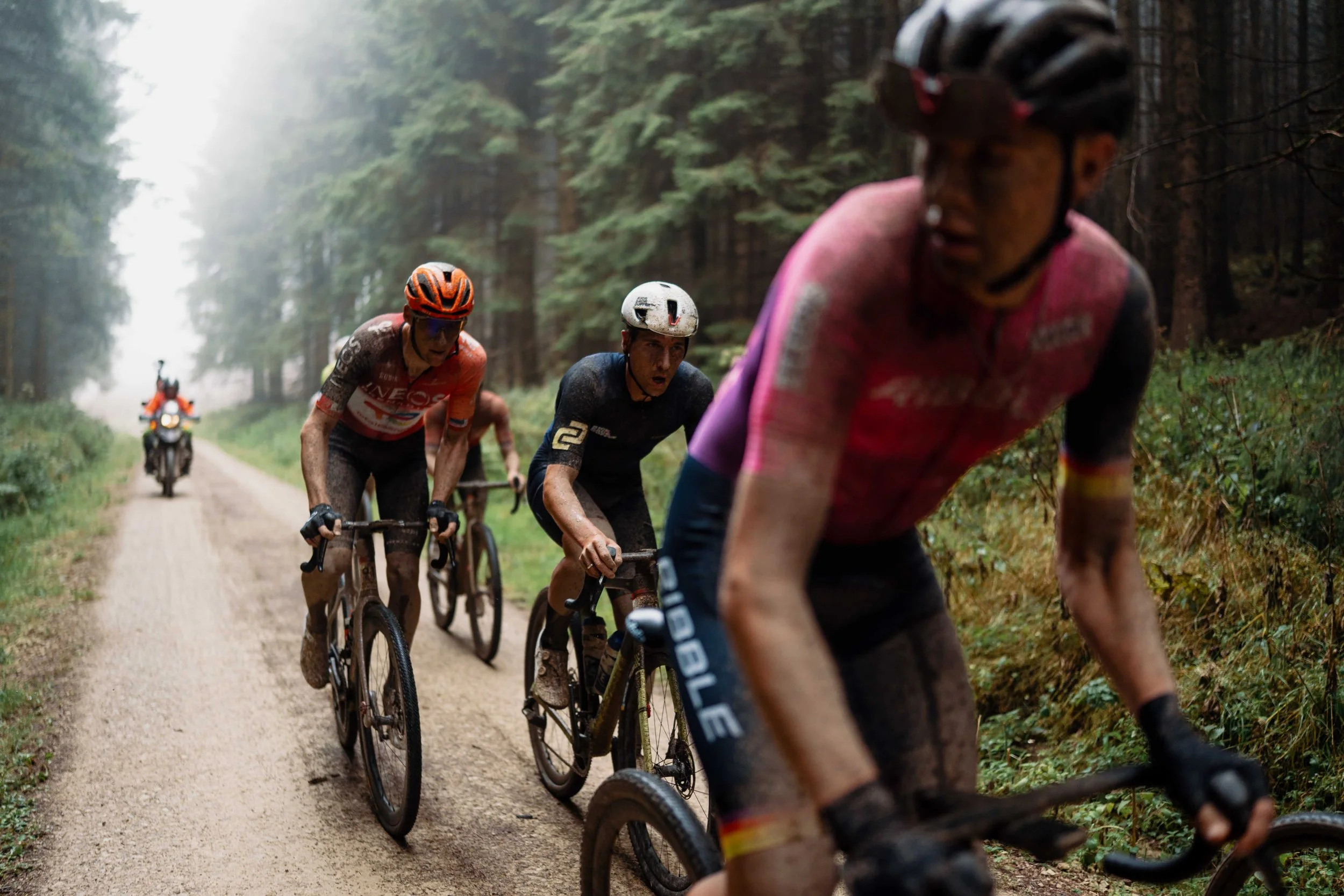 Cyclists riding mountain bikes on a dirt trail through a forest, dirt and mud on their clothing and bikes, with a motorcycle following behind.