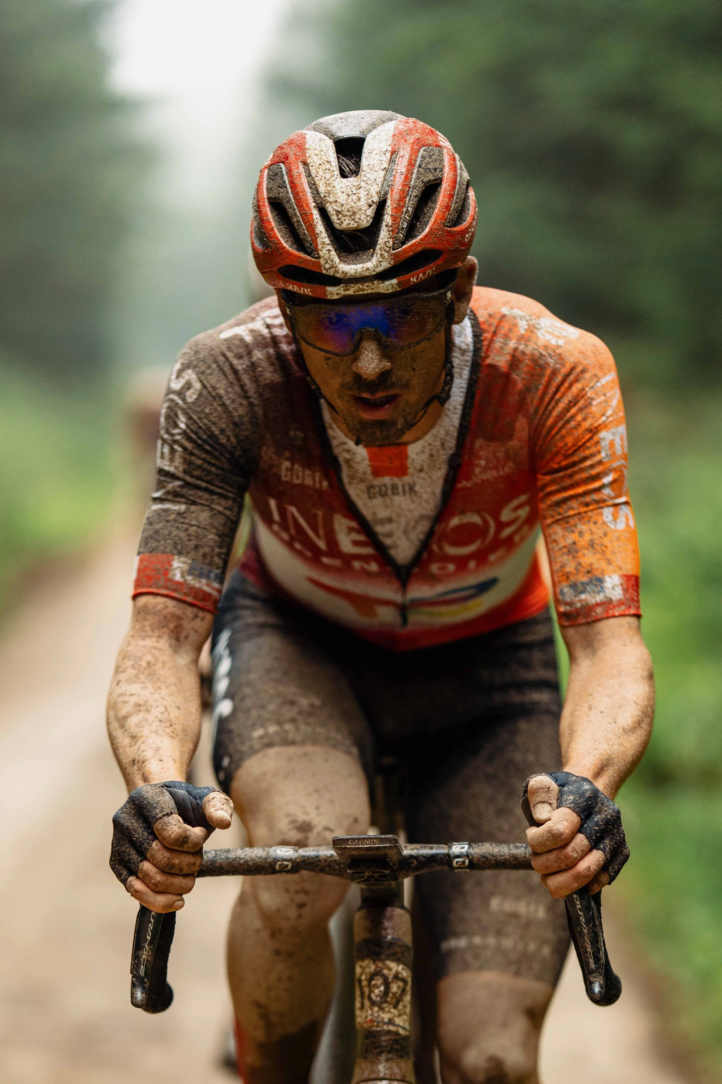 A male cyclist wearing a red and white helmet and sunglasses, riding a muddy bike through a forest trail.